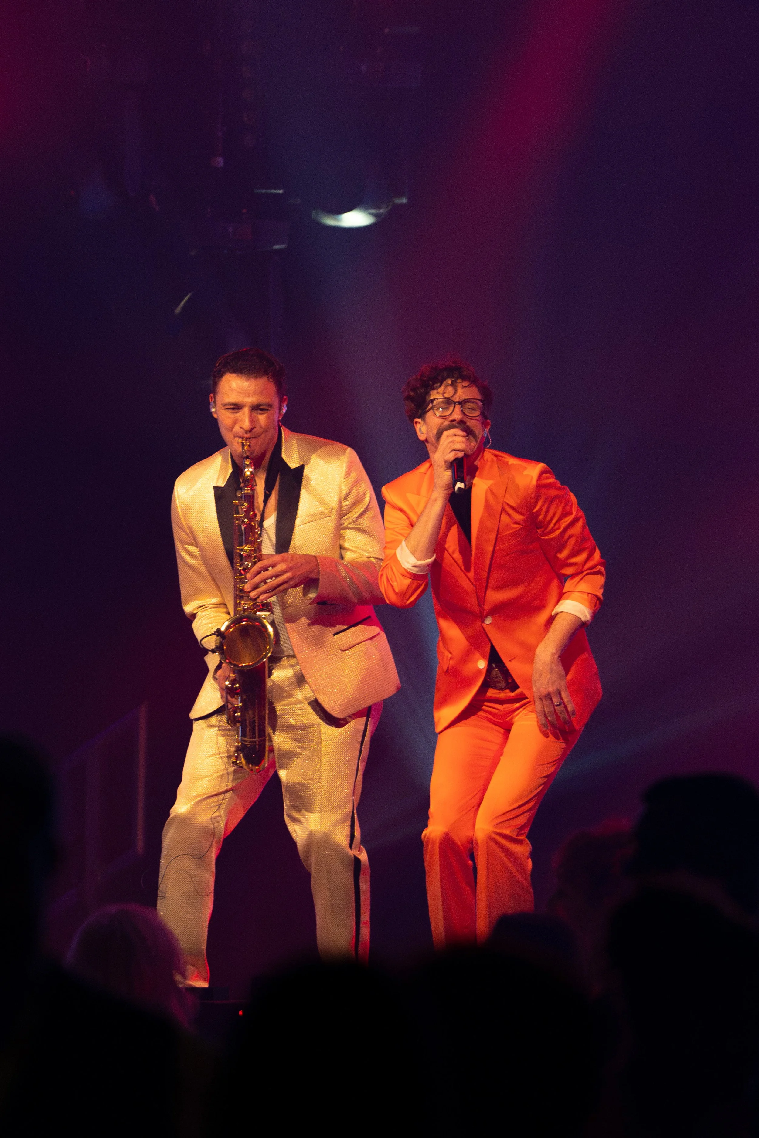Two male performers on stage, one playing a saxophone and the other singing into a microphone, dressed in colorful suits, with a dark background and stage lighting.