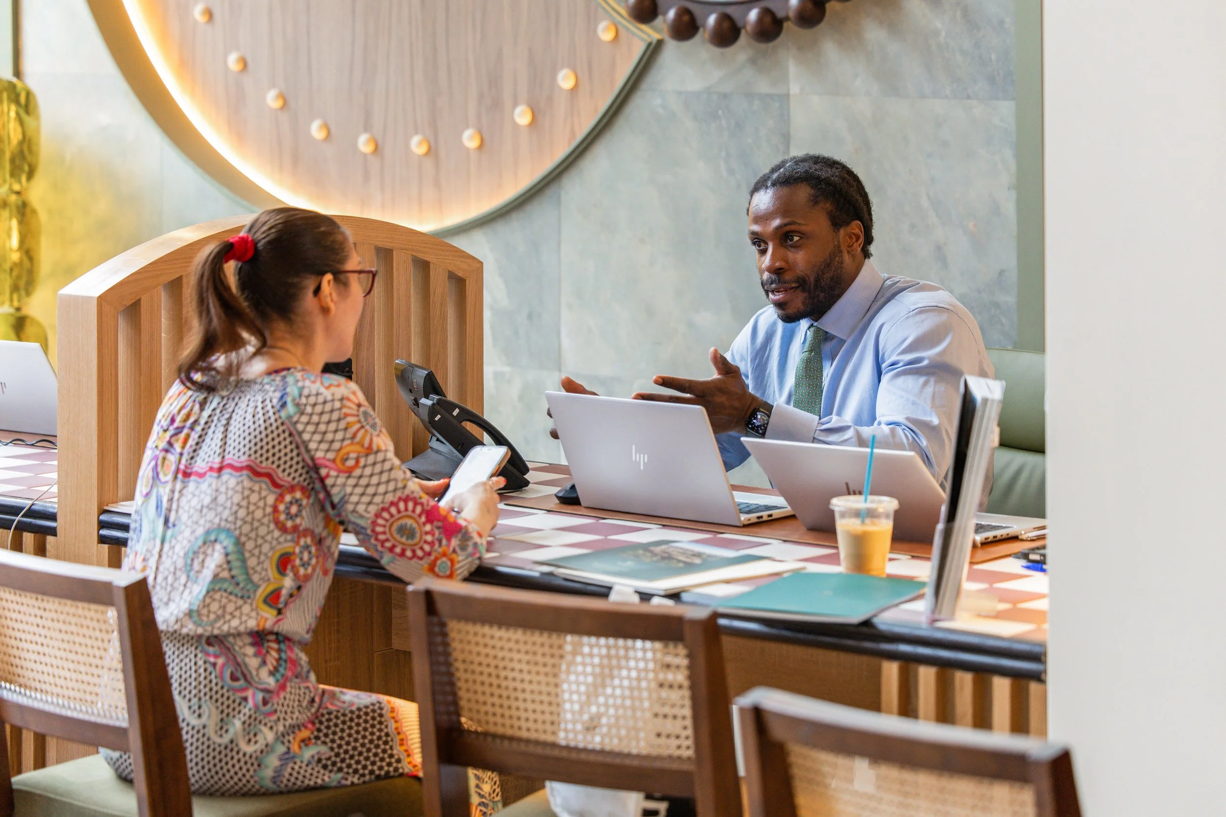 A woman with glasses and a colorful patterned dress sits at a table, engaging in conversation with a man in a light blue shirt and green tie, who is gesturing with his hand. They are in a modern, well-lit cafe or restaurant with wooden accents and a decorative wall in the background. On the table are laptops, a smartphone, and a yellow drink.