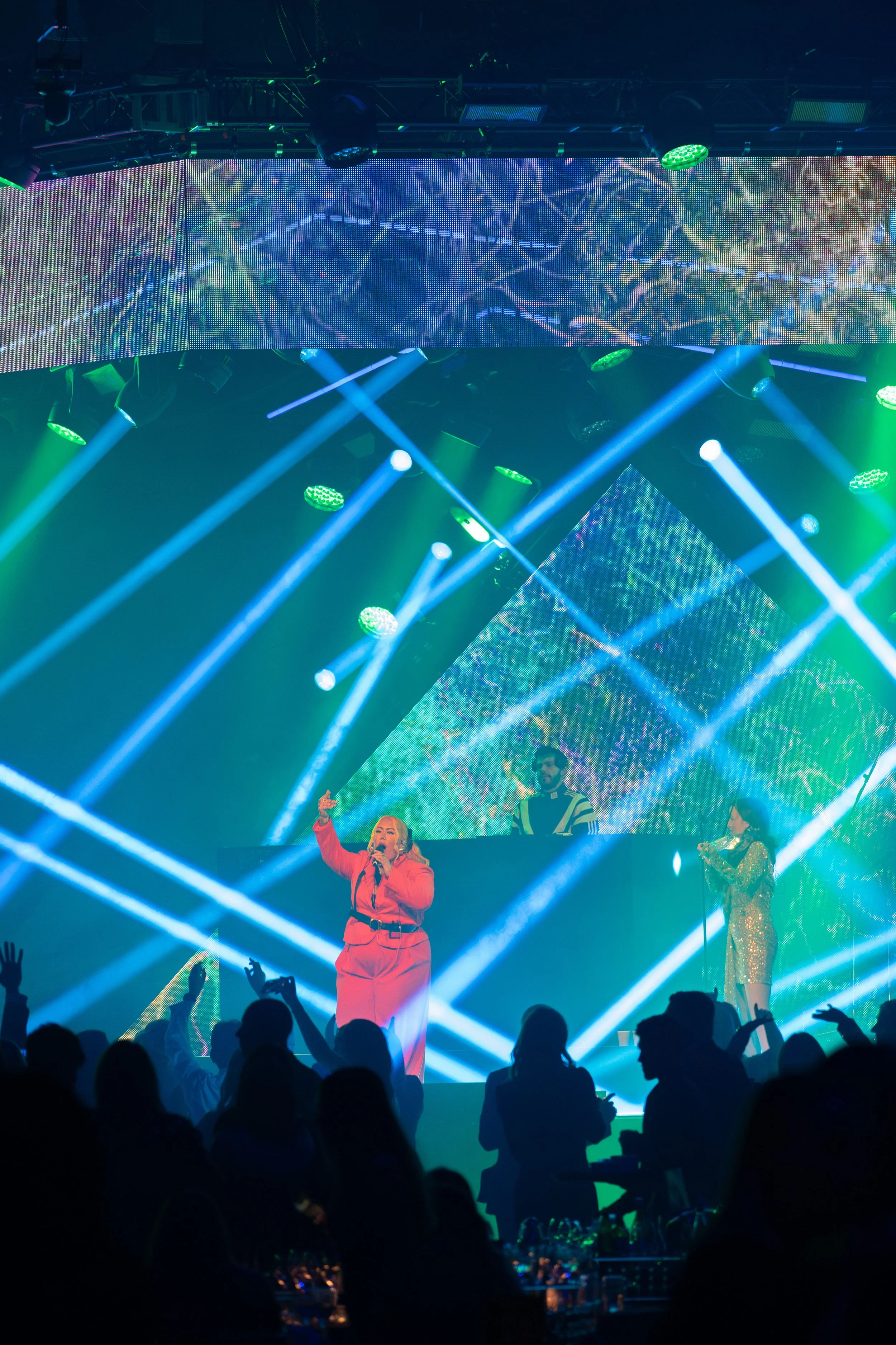 A concert scene with a stage illuminated by blue, green, and white lights, and large screens displaying colorful abstract visuals. A female singer in a red outfit is performing at the front, with her hand raised. Behind her, musicians or performers are visible on the stage. The audience is silhouetted in the foreground, watching and enjoying the performance.
