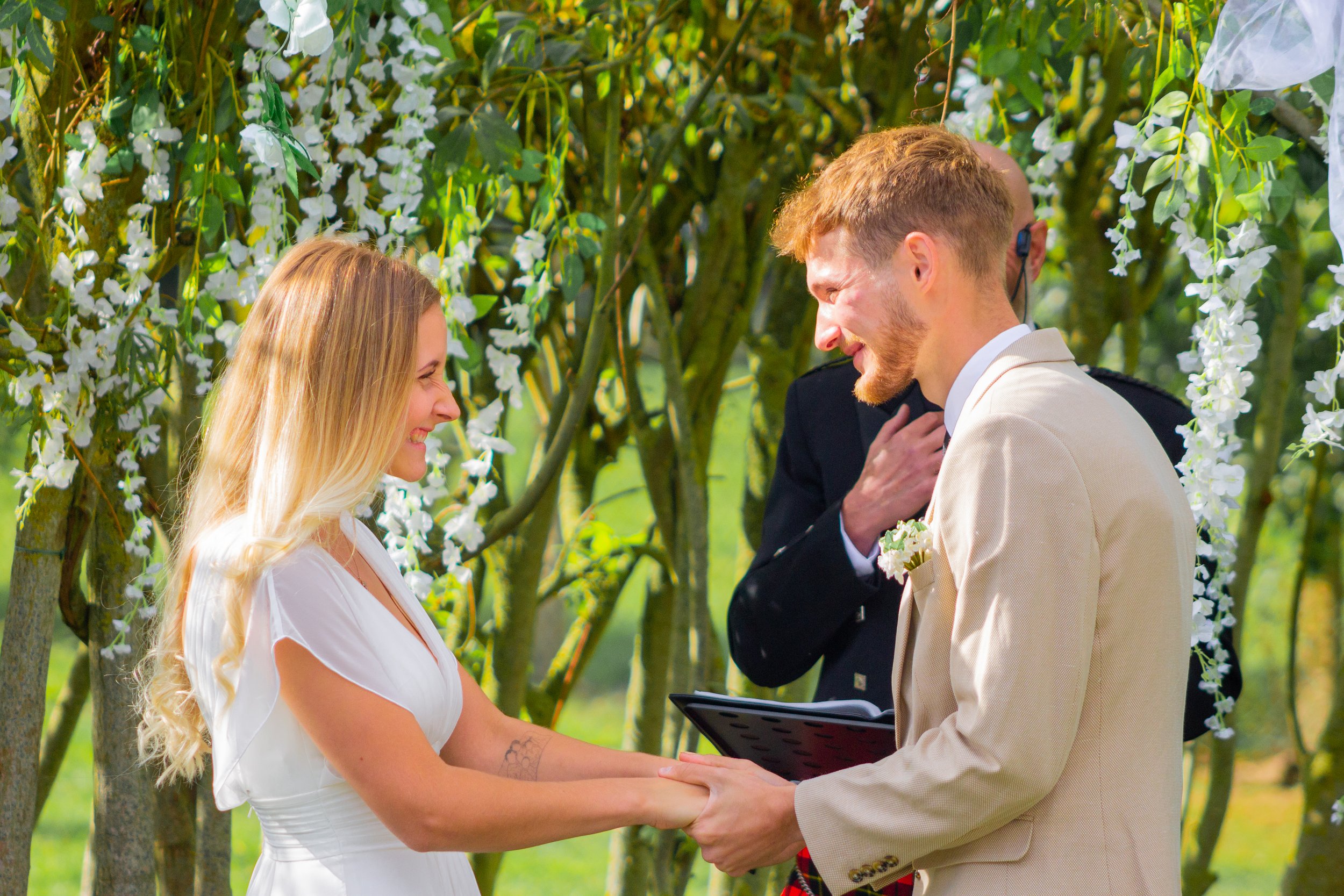 A couple getting married outdoors with a person officiating, surrounded by trees with white flowers.