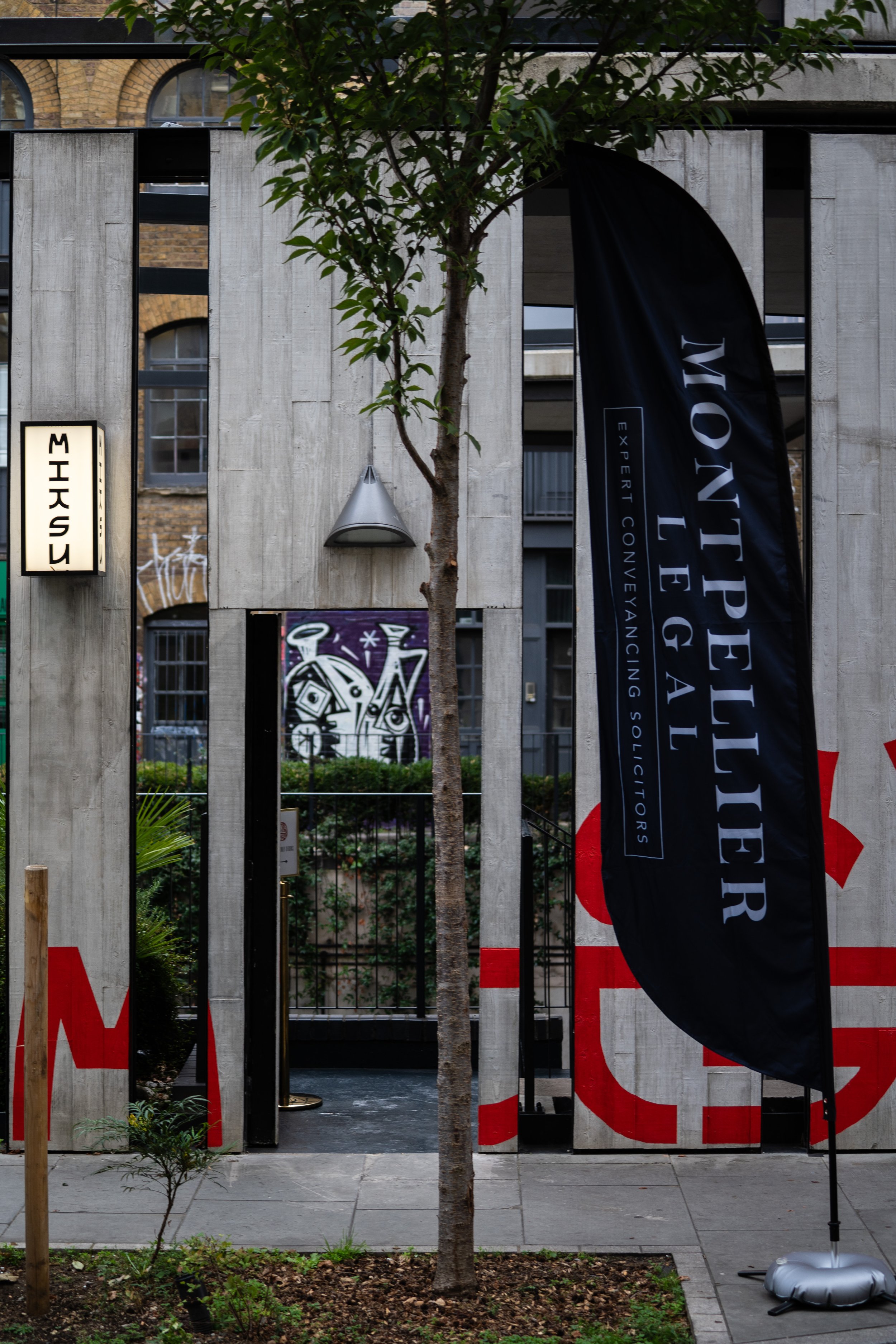 Urban street scene with a tree in the foreground, wooden and brick buildings in the background, a black Flyer flag with the text 'Montpellier' and additional phrases, a white sign with black text, and various graffiti art visible through a window.