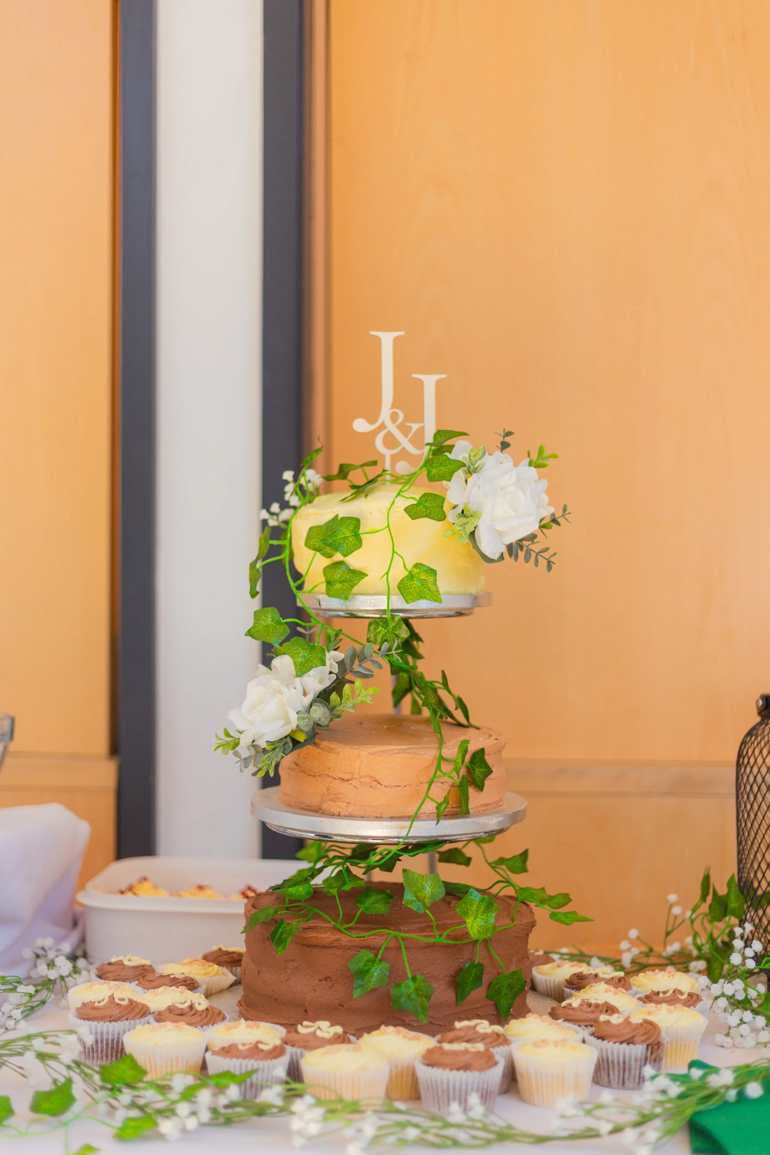 A large tiered cake decorated with green ivy and white flowers, positioned on a round wooden stand. Surrounding the cake are cups of assorted cupcakes with swirled frosting. The background includes wooden panels and a table with additional desserts.