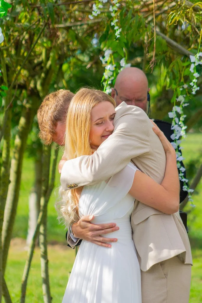 A woman in a white dress and two men hugging in an outdoor garden setting, surrounded by greenery and hanging flowers.