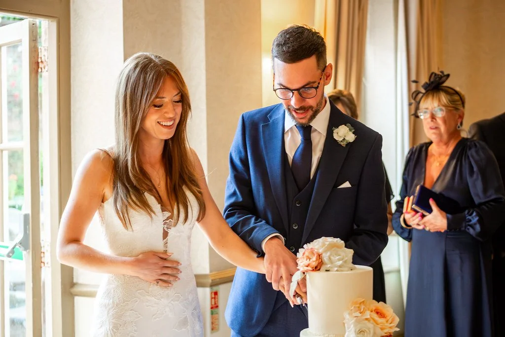 A bride and groom cutting a wedding cake, with a woman in formal attire watching in the background.