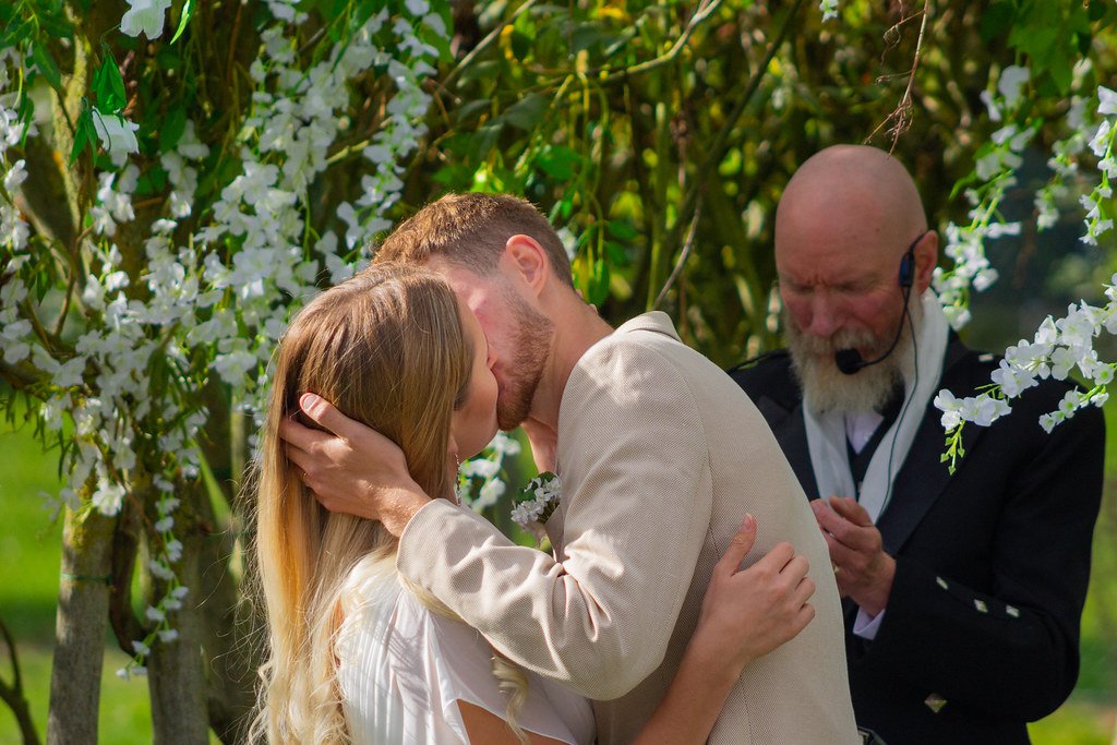 A couple kissing during their outdoor wedding ceremony under a flowering tree, with an officiant or priest in a black suit overseeing the ceremony.