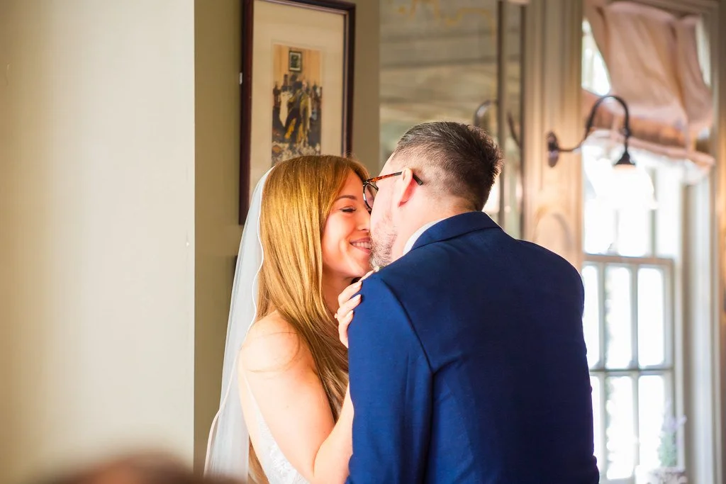 A bride with long red hair and a white wedding dress smiling as she leans into a groom in a dark blue suit, sharing an intimate moment indoors near windows with curtains.