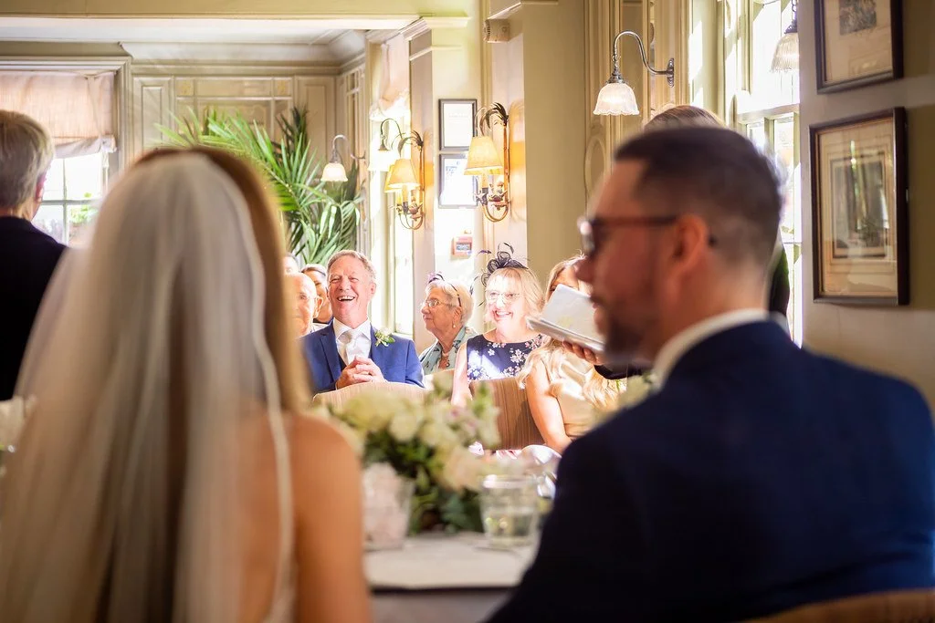 A bride and groom are seated at a table during their wedding reception, with guests in the background enjoying the celebration in a cozy, well-lit room decorated with plants and artwork.