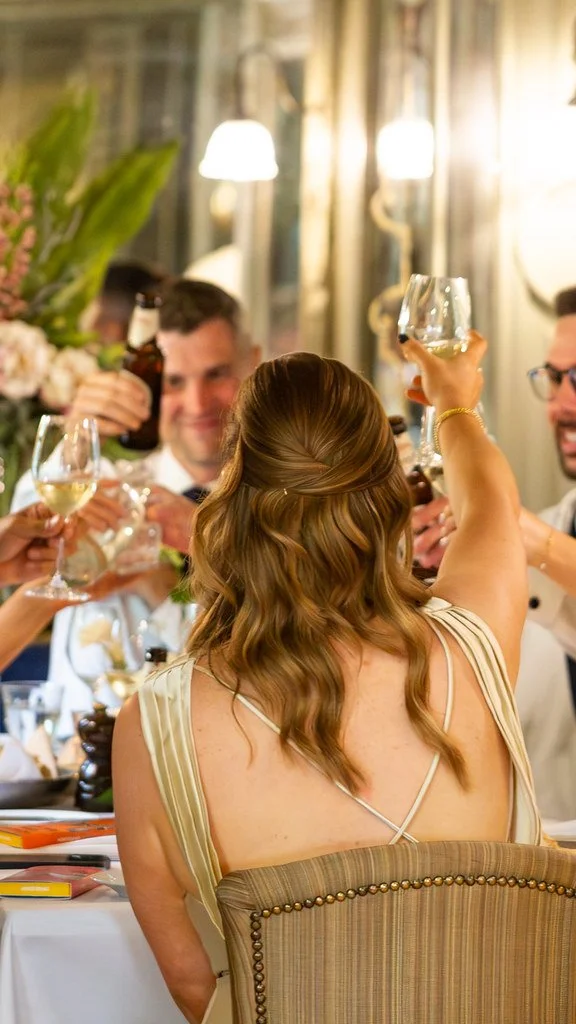 People celebrating at a dinner party with raised glasses, including a woman with wavy hair and a sleeveless dress at a decorated table.