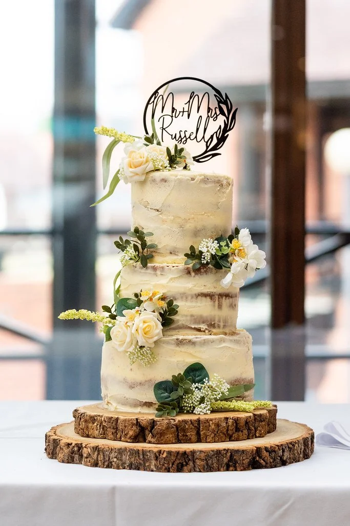 Three-tier semi-naked wedding cake decorated with white roses, greenery, and small white flowers, placed on a rustic wooden cake stand with a black personalized cake topper reading "Mr & Mrs Russell" in a circle design.