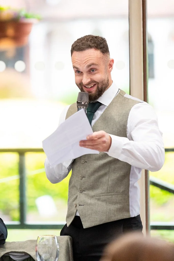 A man in a beige vest, white shirt, and dark tie smiling and speaking into a microphone while holding a paper.