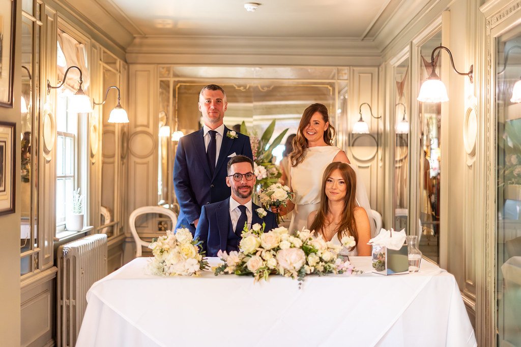 Four people at a wedding reception, two men and two women, with the bride holding a bouquet, sitting and standing behind a table decorated with white flowers.