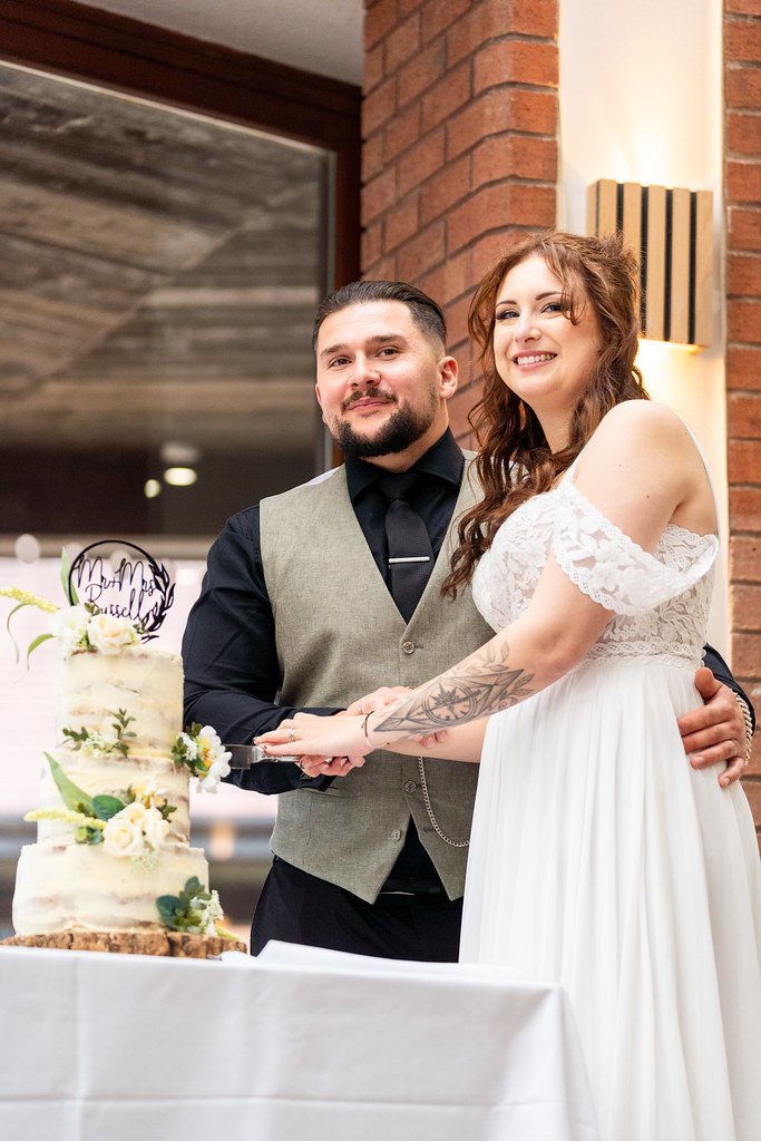 A newlywed couple cuts their wedding cake together. The bride wears a white lace wedding dress, and the groom wears a black shirt with a grey vest. They are smiling and holding a cake knife with their hands together.