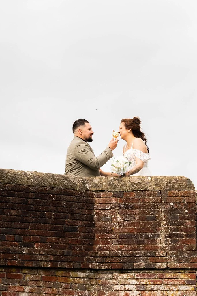 A man in a beige blazer and a woman in a white dress sharing a piece of cake on top of a brick wall, with the woman holding a bouquet of white flowers, against a gray sky background.