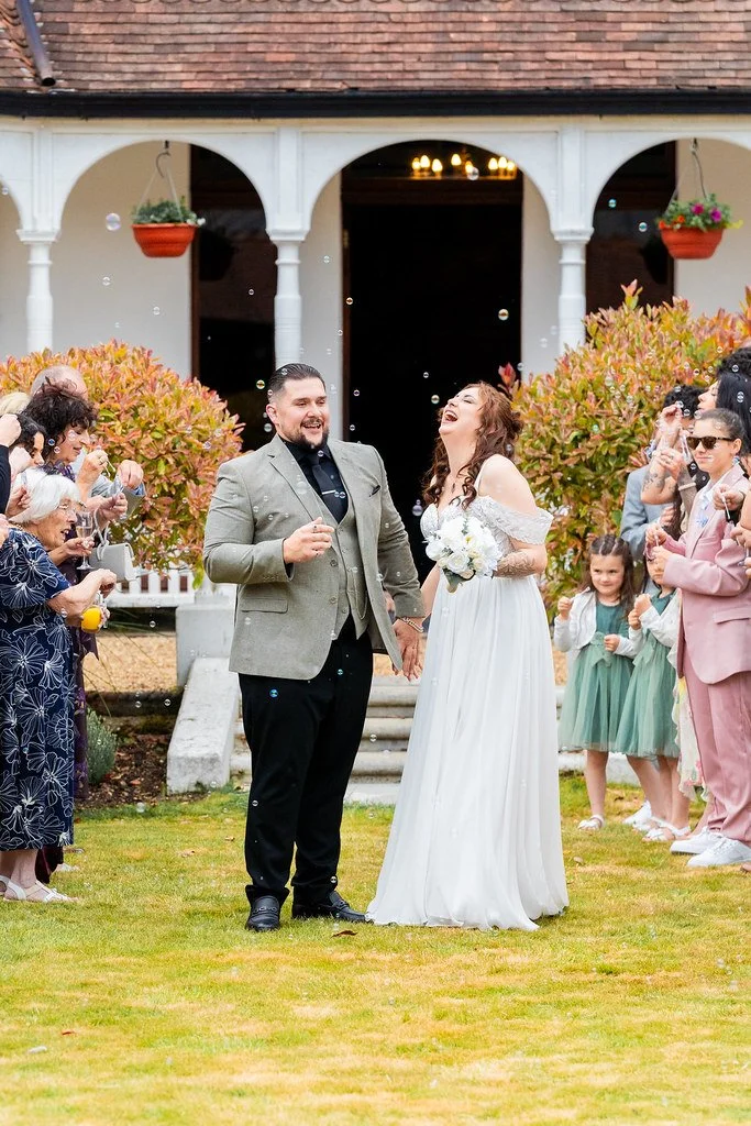 A bride and groom holding hands and smiling, celebrating their wedding outdoors with friends and family playing with bubbles around them.