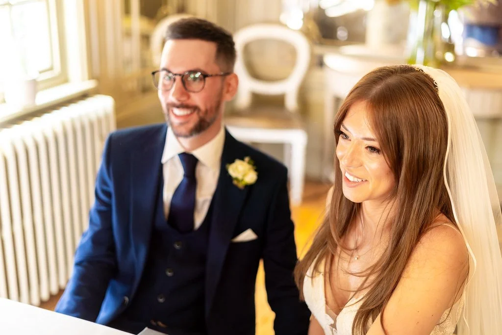 A bride and groom smiling at their wedding ceremony, seated indoors with warm lighting, the groom wearing glasses and a navy suit with a boutonniere, the bride wearing a white dress and veil.