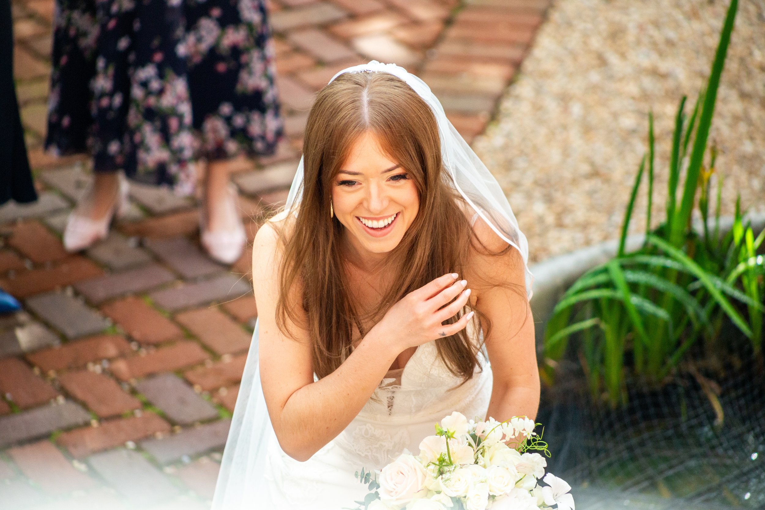 A smiling woman in a white dress, likely a bride, holding a bouquet of white flowers, with a brick walkway, greenery, and another person in a dark floral dress in the background.