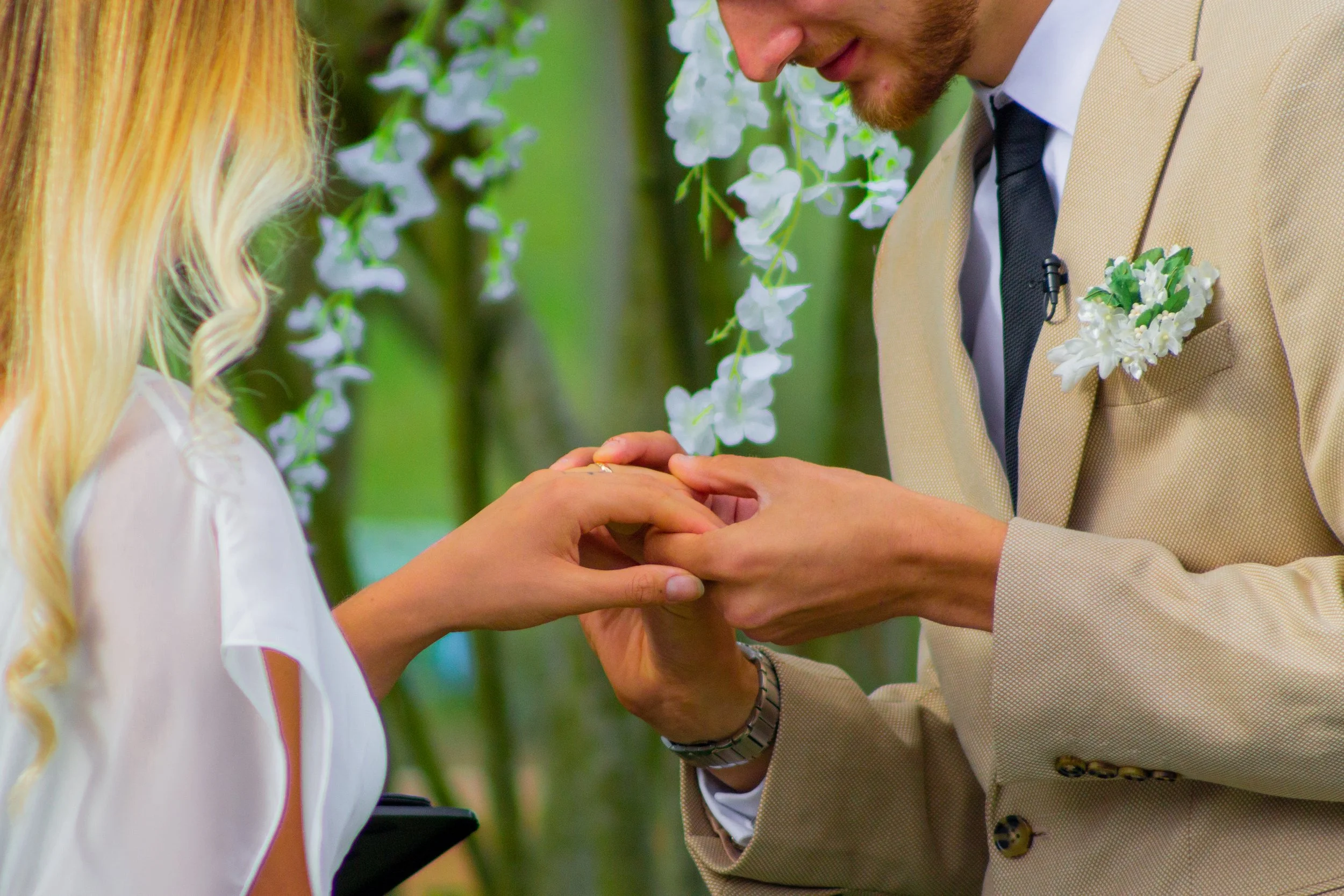 Man in a tan suit exchanges rings with a woman during a wedding ceremony outside, with white flowers and green foliage in the background.