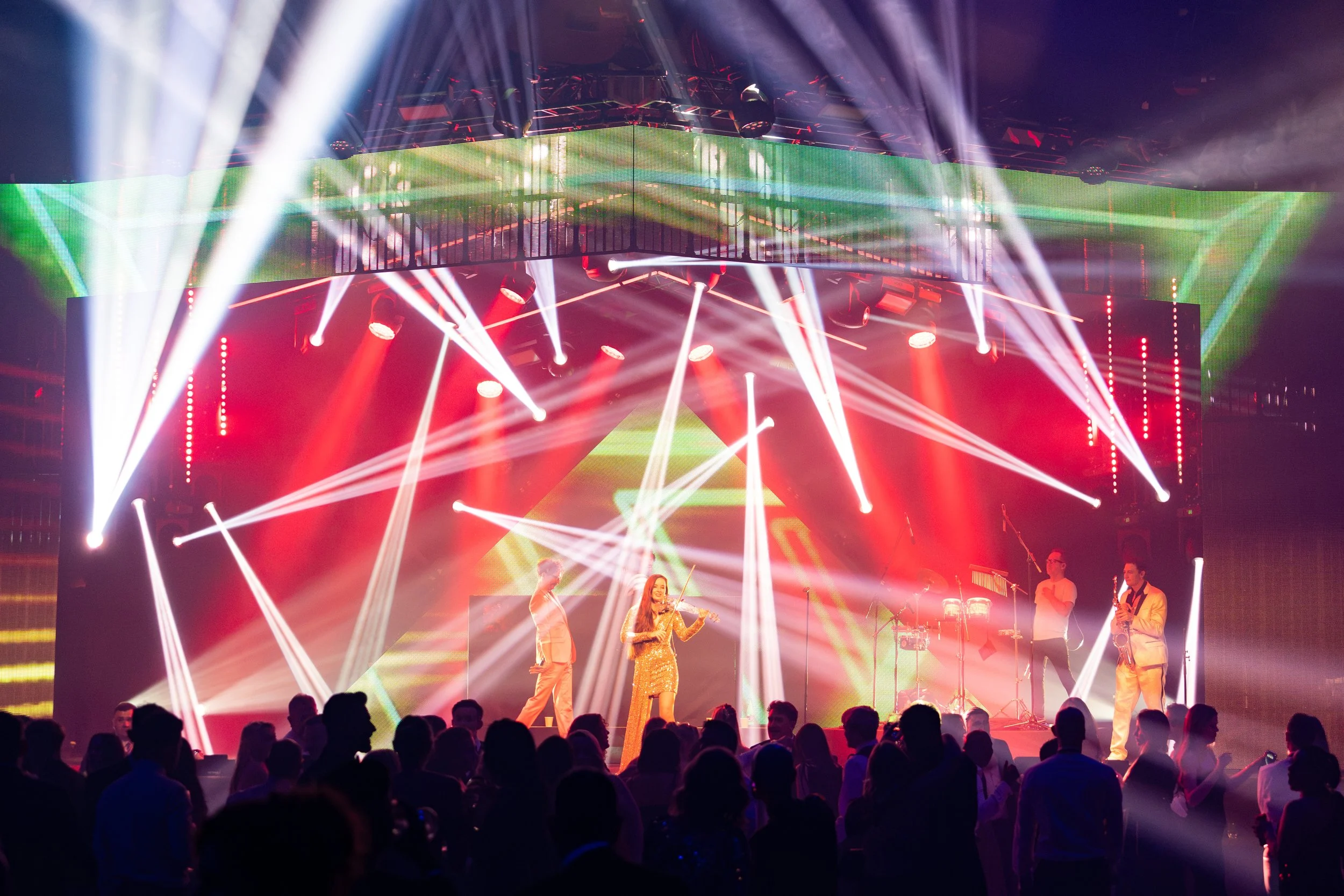 A live music concert features performers on stage surrounded by vibrant red, green, and white lighting effects, with a large audience watching.