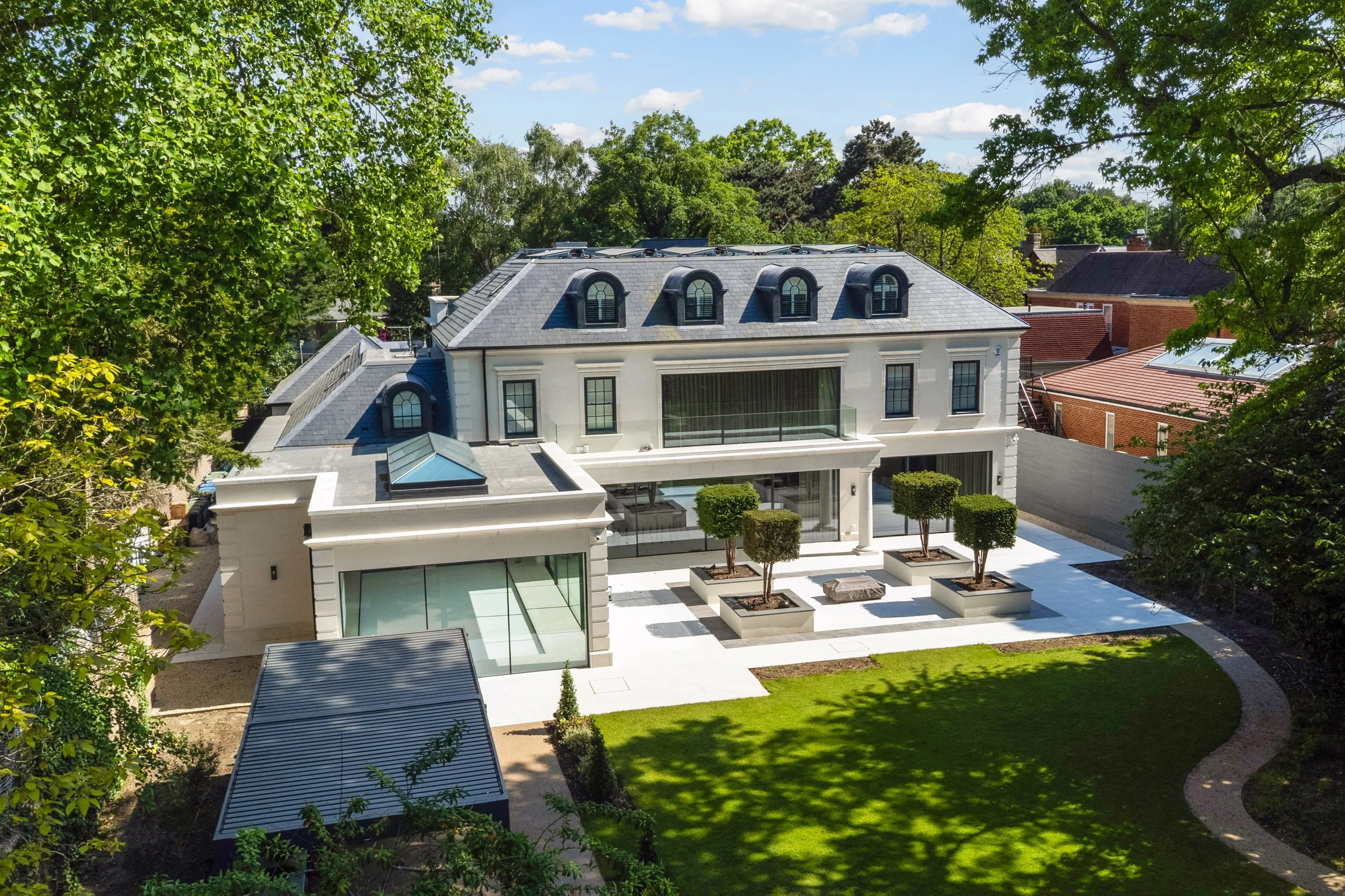 A modern white house with black-framed windows and a gray slate roof, surrounded by a landscaped yard with small trees and manicured lawn, partially shaded by green trees in the foreground, under a partly cloudy blue sky.