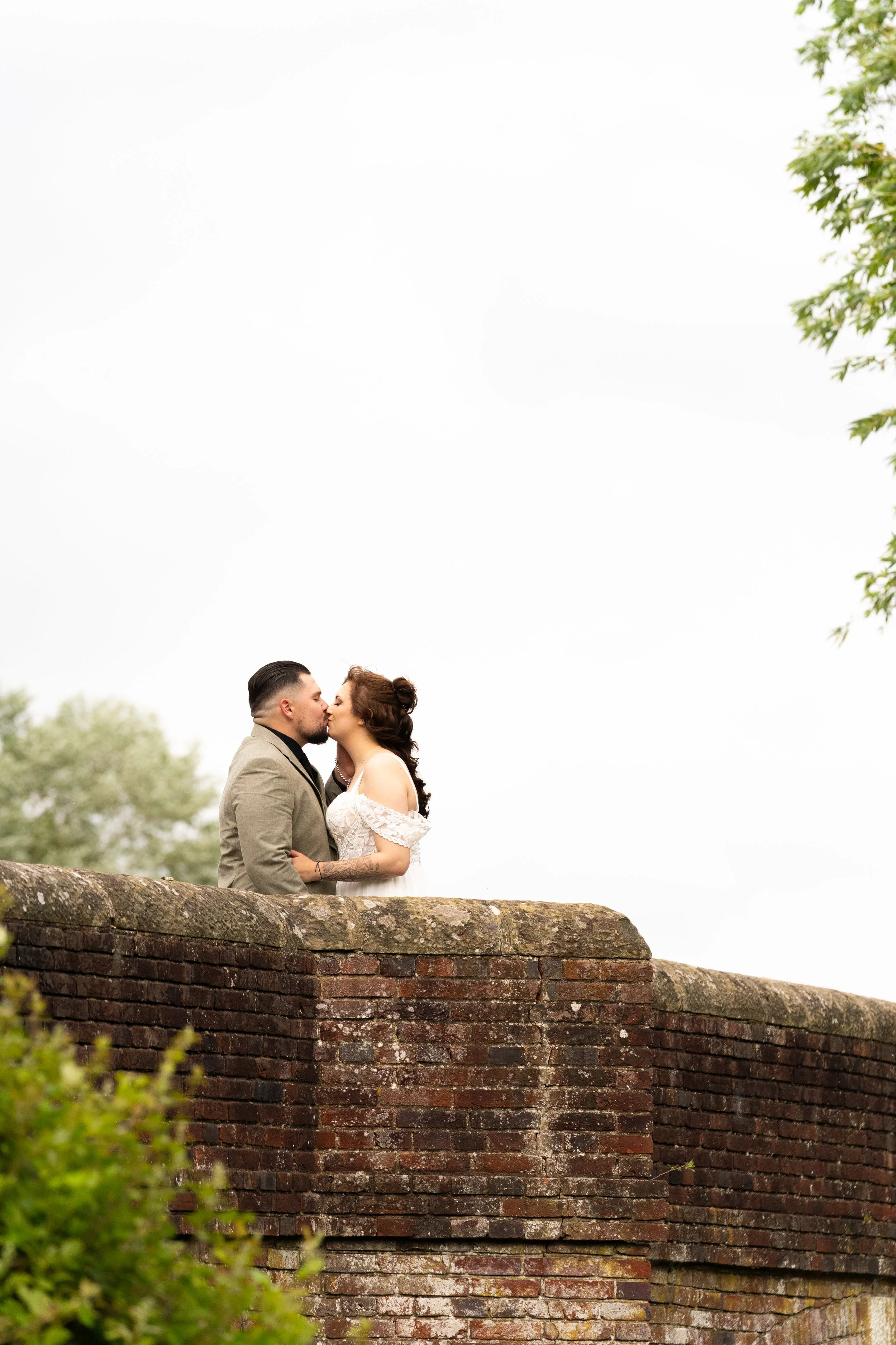 A couple dressed in wedding attire sharing a kiss outdoors, standing behind an old brick wall with green foliage in the foreground and trees in the background.