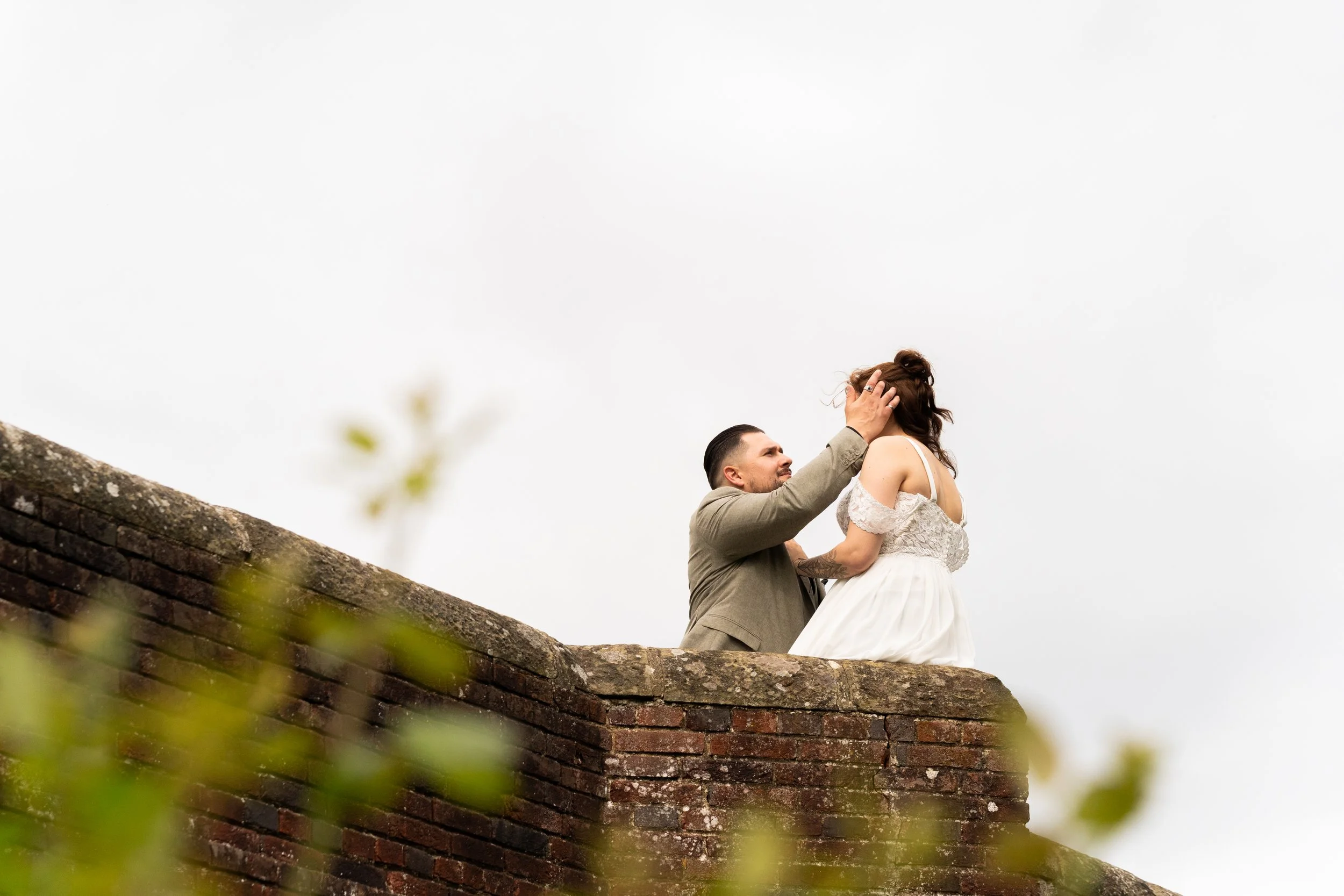 A man in a beige suit lifting a woman in a white dress while kneeling on a brick wall outdoors with a cloudy sky in the background.