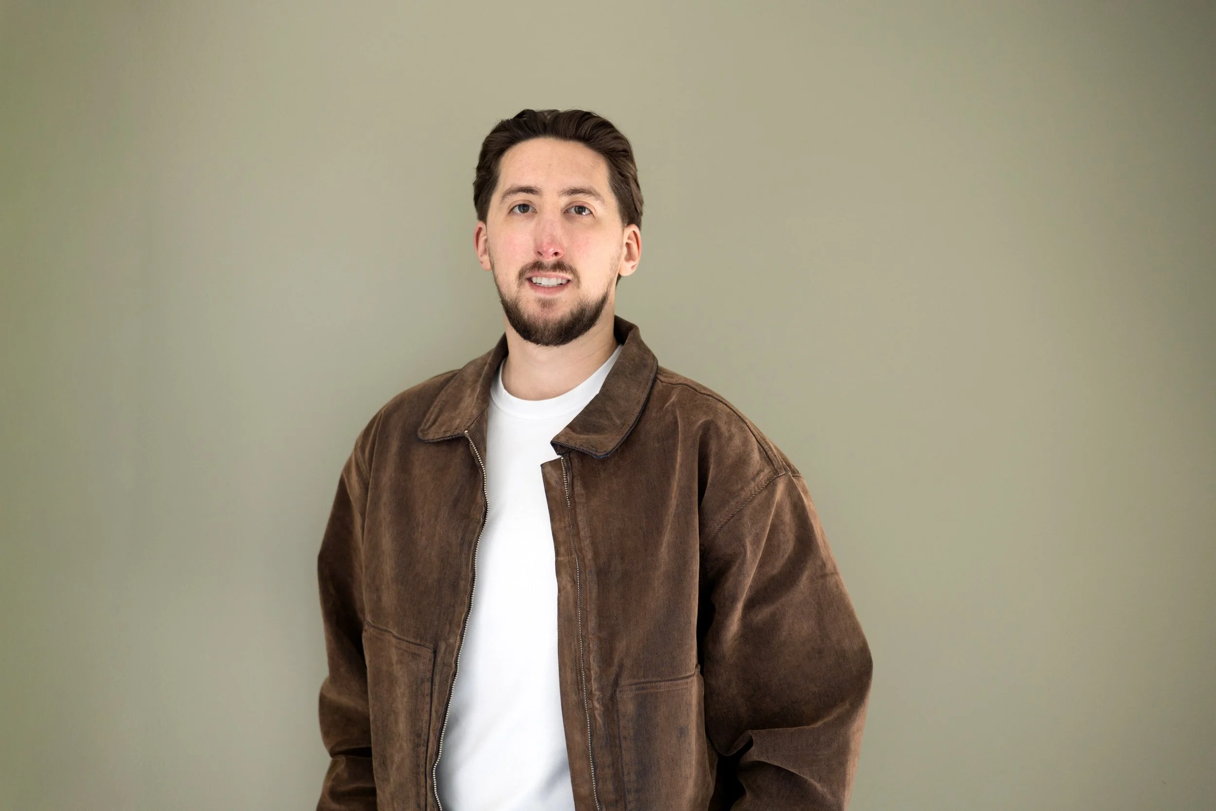 A young man with brown hair, beard, and mustache, wearing a brown jacket over a white t-shirt, standing against a plain light-colored wall.