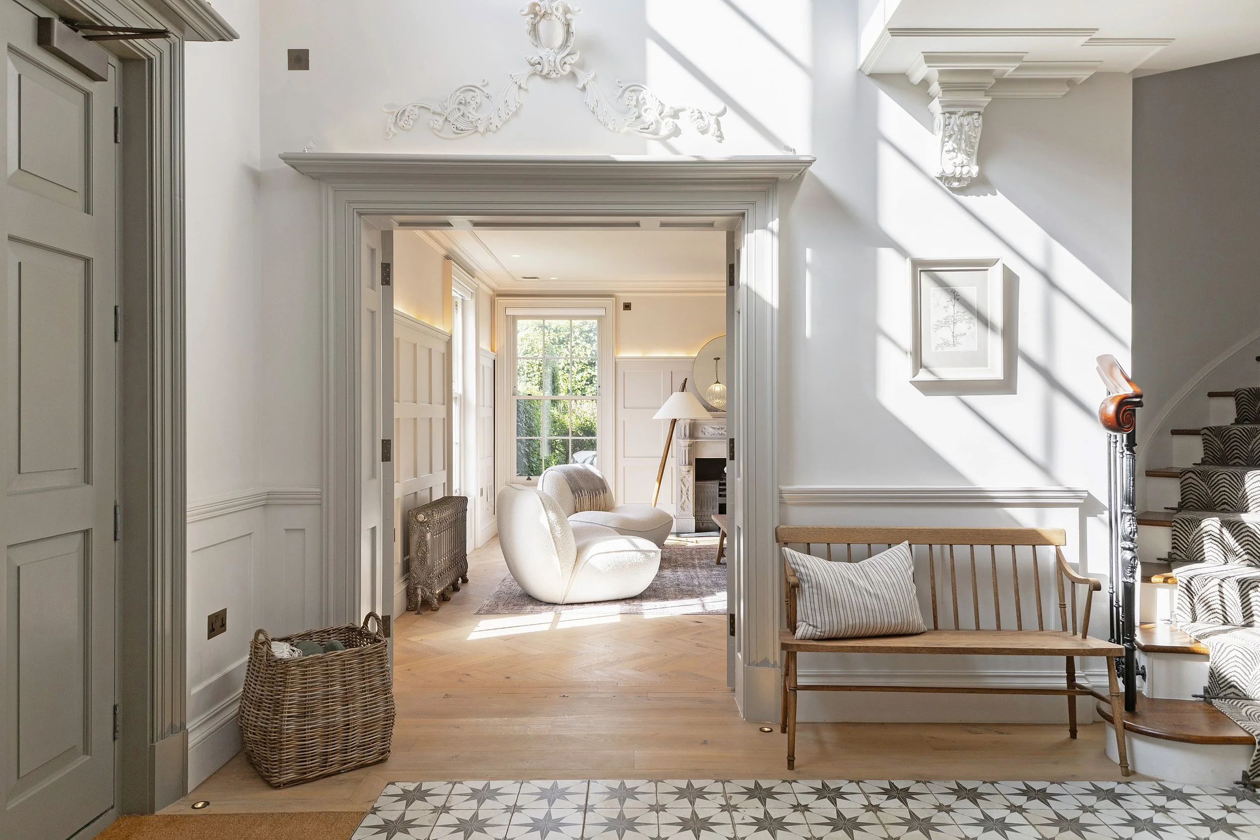 Bright and airy living room with large window, white walls, and ornate crown molding. Includes white armchairs, a fireplace, a standing lamp, and decorative framed artwork.