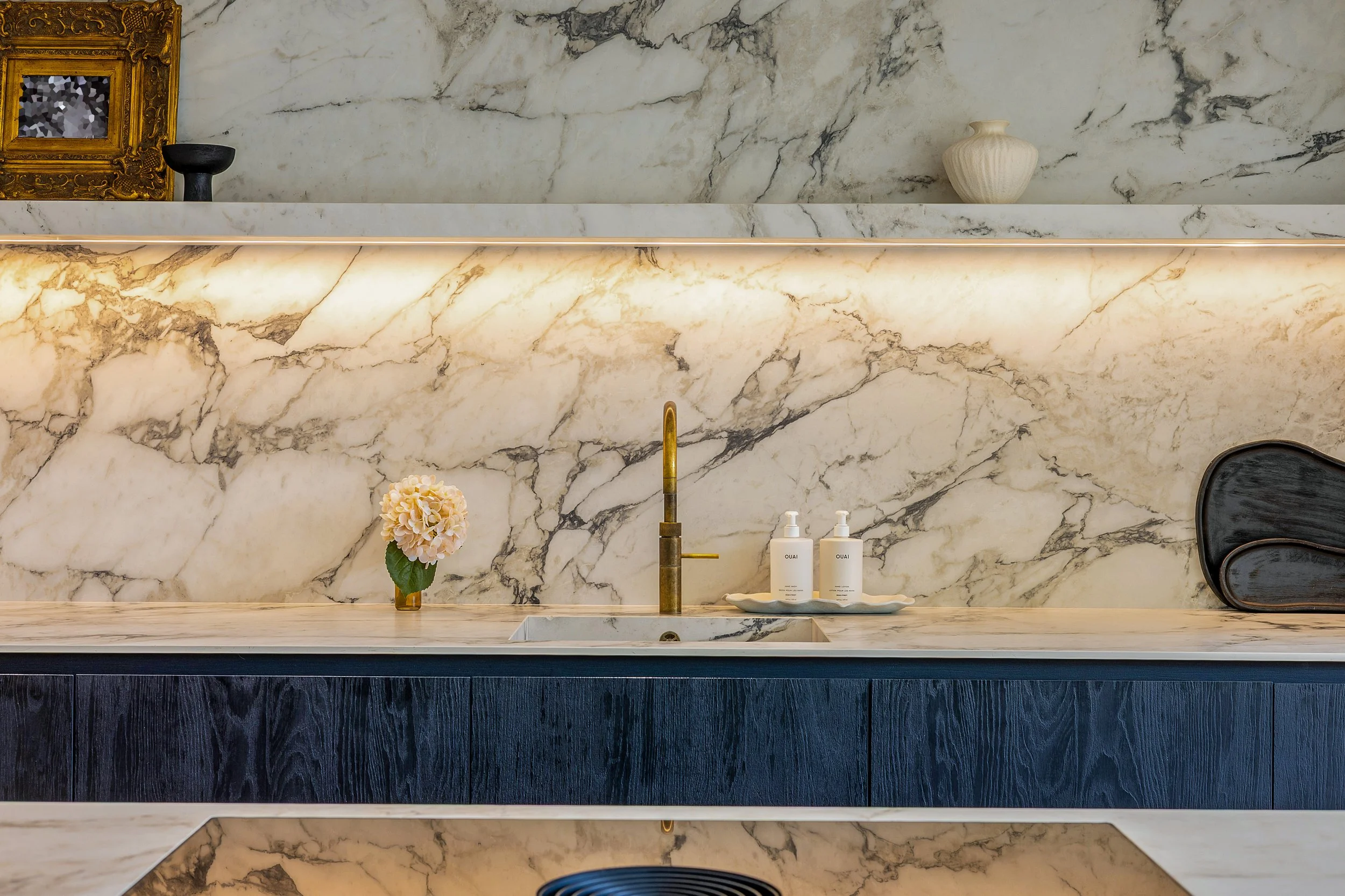 A modern bathroom with a marble backsplash and countertop. The countertop features a gold faucet, a small vase with pink flowers, and two white bottles labeled 'OUAI.' There are decorative objects on the marble shelf, including a black bowl, a black chair, and a plain white vase.