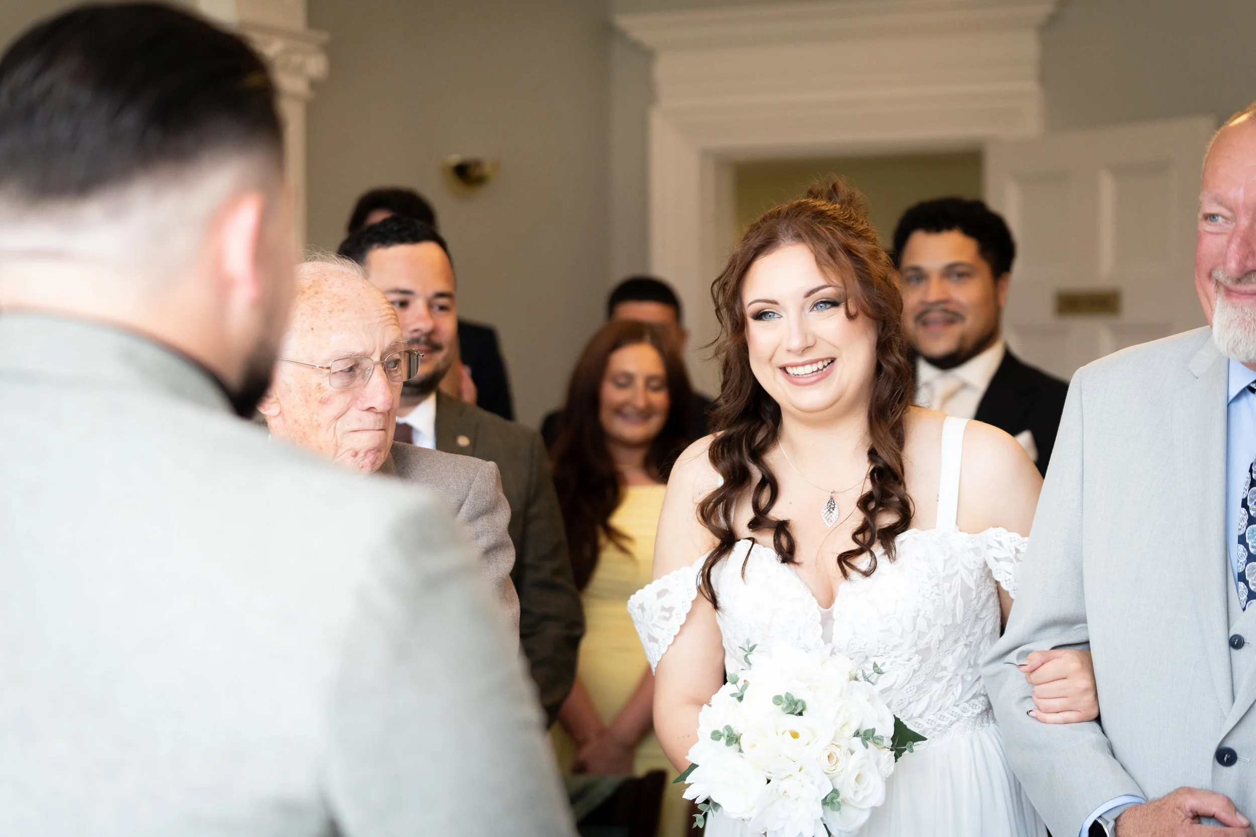 A bride with long, curly brown hair wearing a white dress and holding a bouquet of white roses and greenery, smiling during a wedding ceremony with several people dressed formally in the background.
