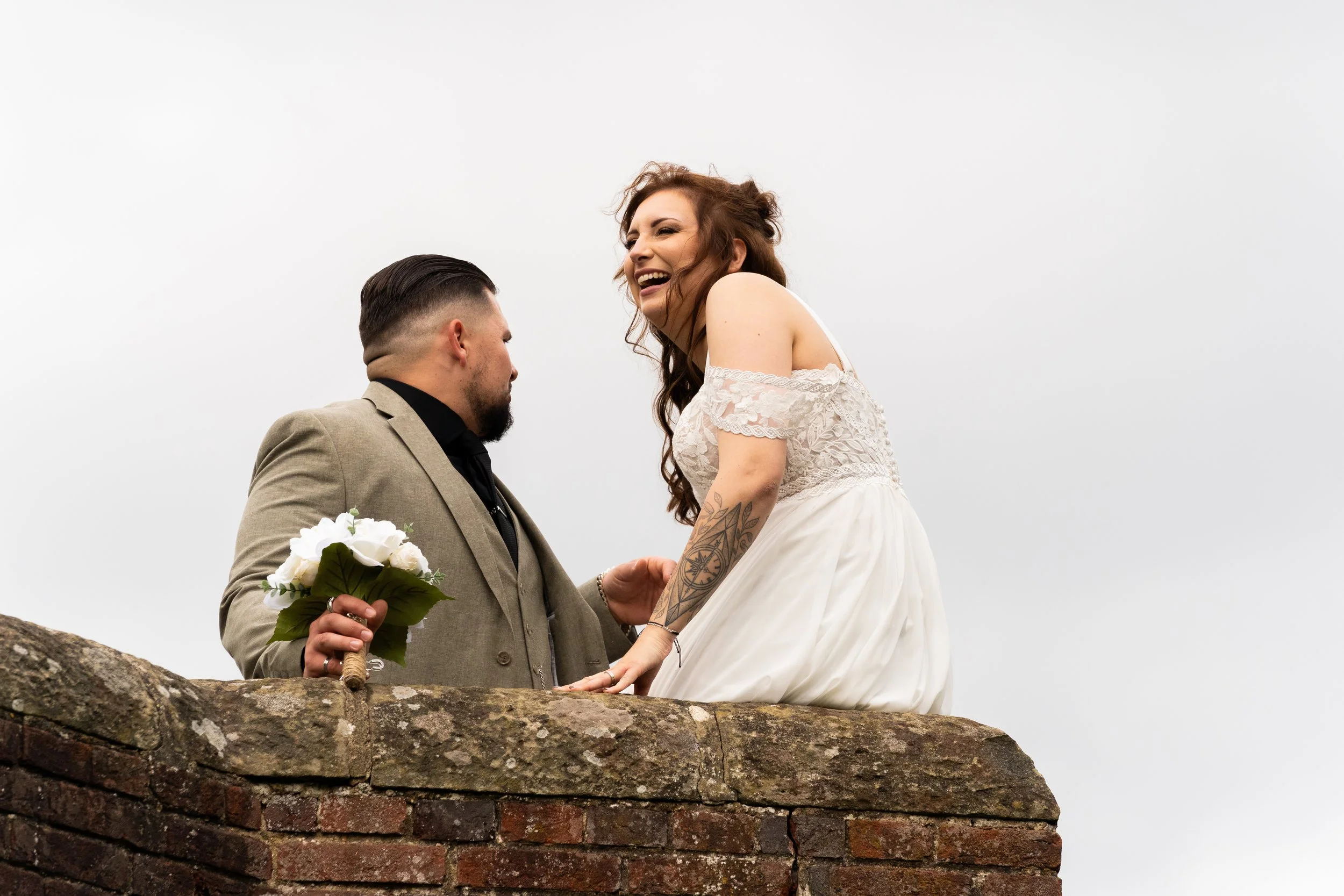 A couple on their wedding day is standing behind an old brick wall outdoors. The groom is wearing a light-colored suit, holding a bouquet of white roses, and is looking up at the bride. The bride is wearing a white lace wedding dress, laughing, and leaning slightly toward the groom, with tattoos visible on her arm and long curly hair.
