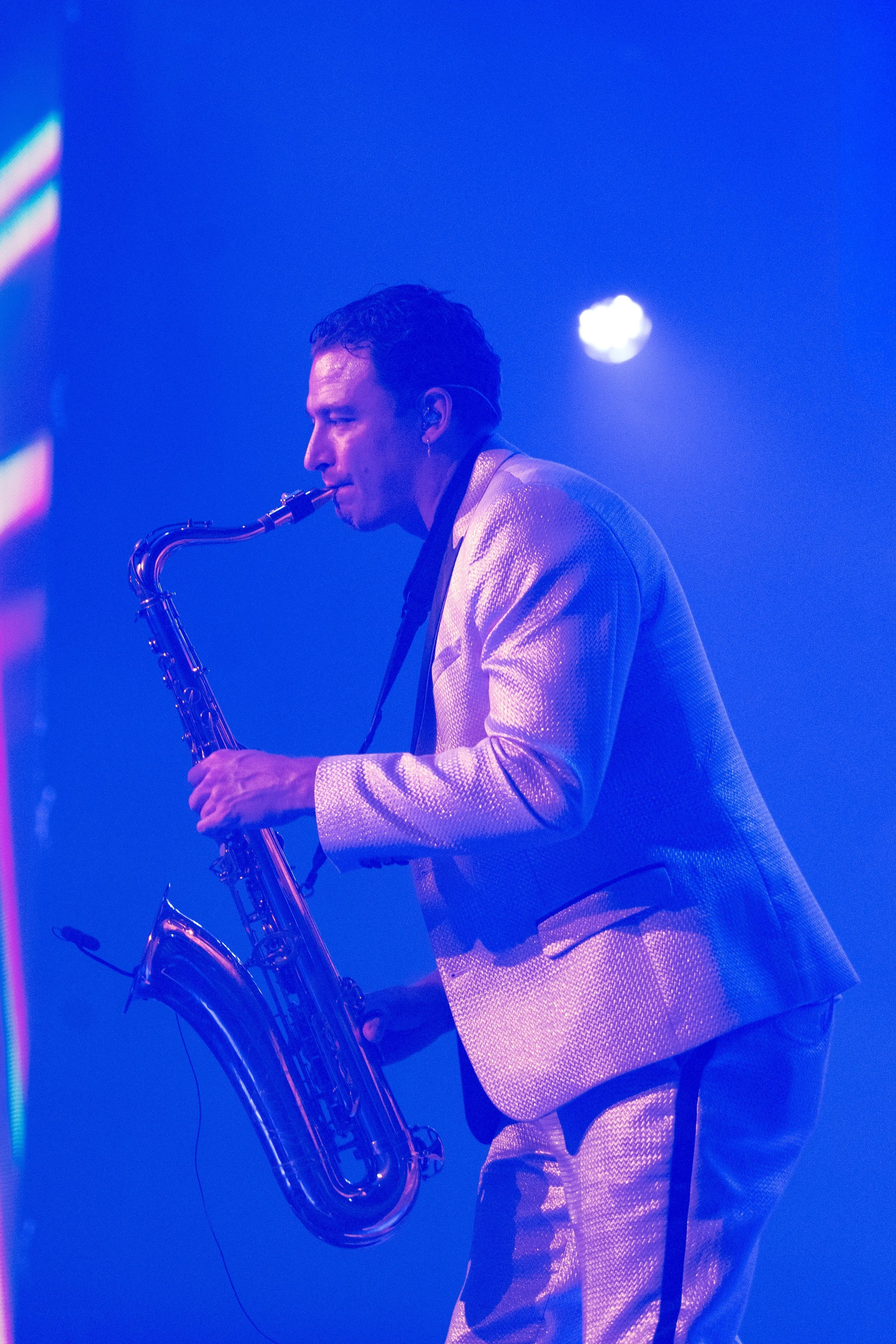 A man playing a saxophone on stage, dressed in a shiny suit, with blue stage lighting and a visible bright light in the background.