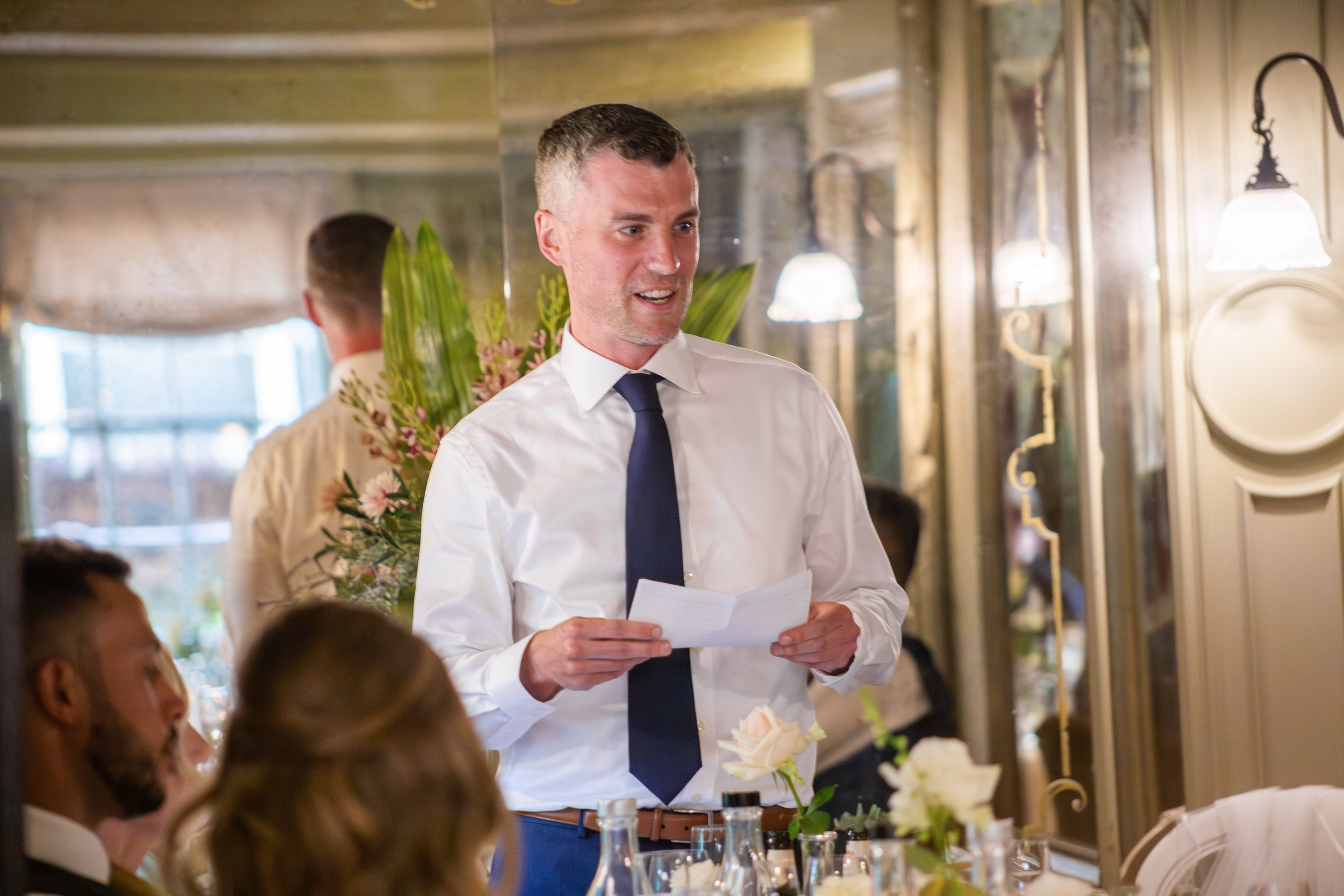 A man in a white shirt and navy tie holding a piece of paper, standing in front of a seated group at a restaurant or banquet with floral decorations and warm lighting.