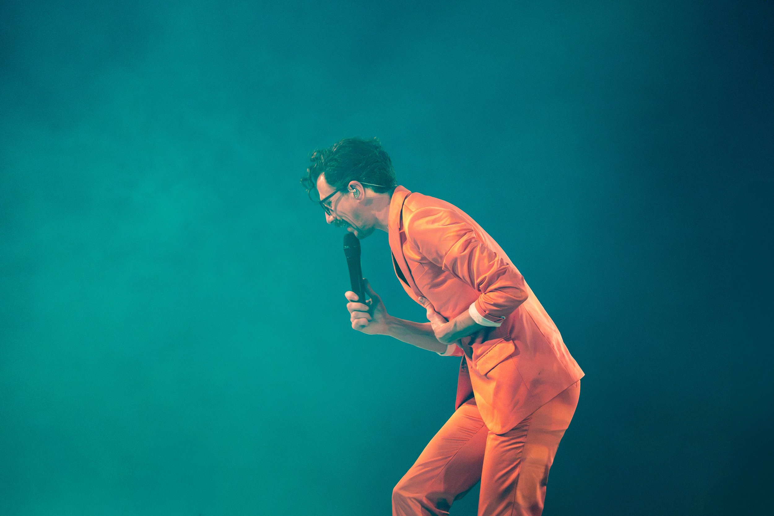 A man with glasses and styled hair wearing an orange outfit, passionately singing into a microphone on stage with a teal background.