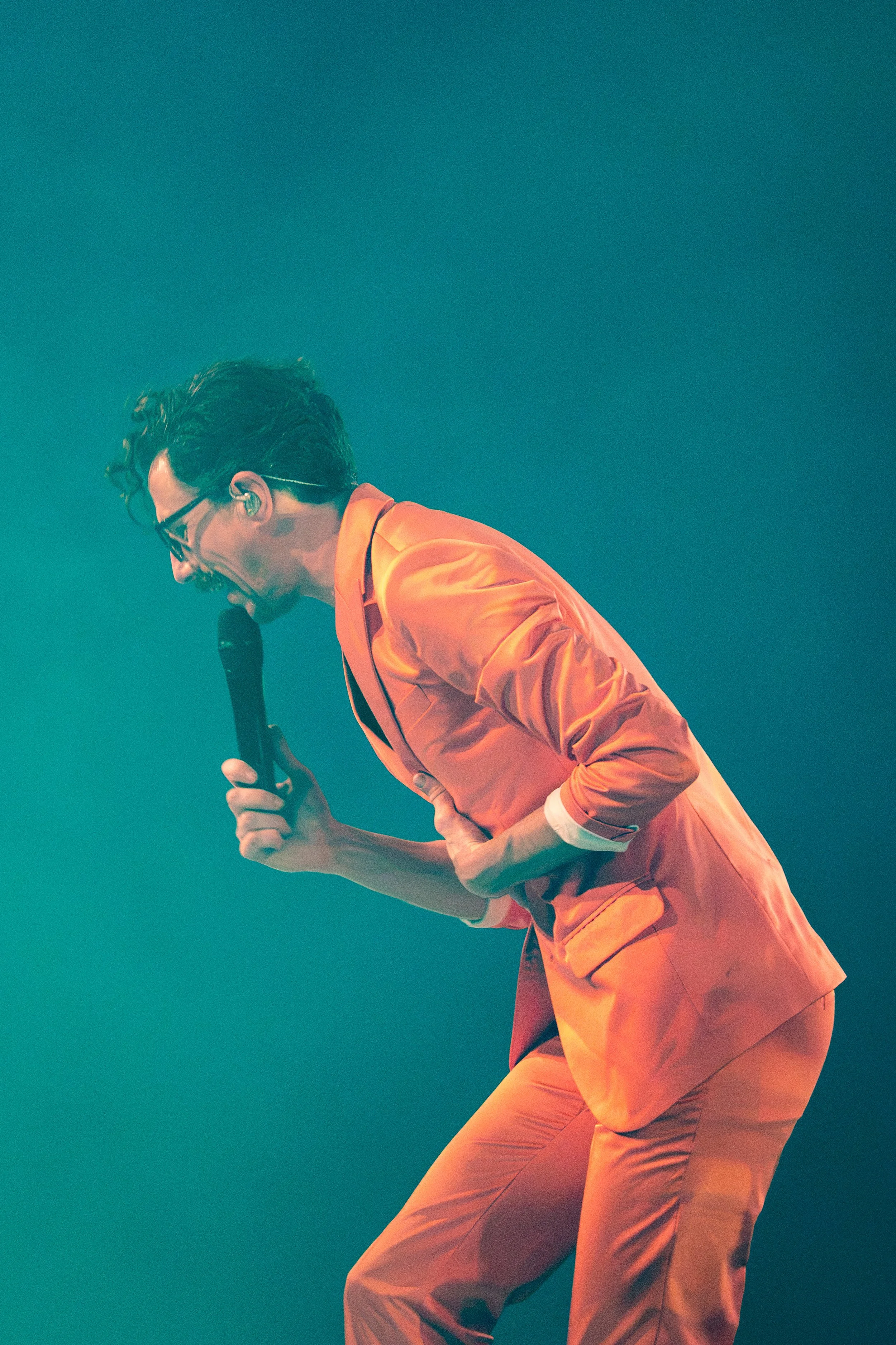 A man in an orange suit with glasses, singing passionately into a microphone against a teal background.