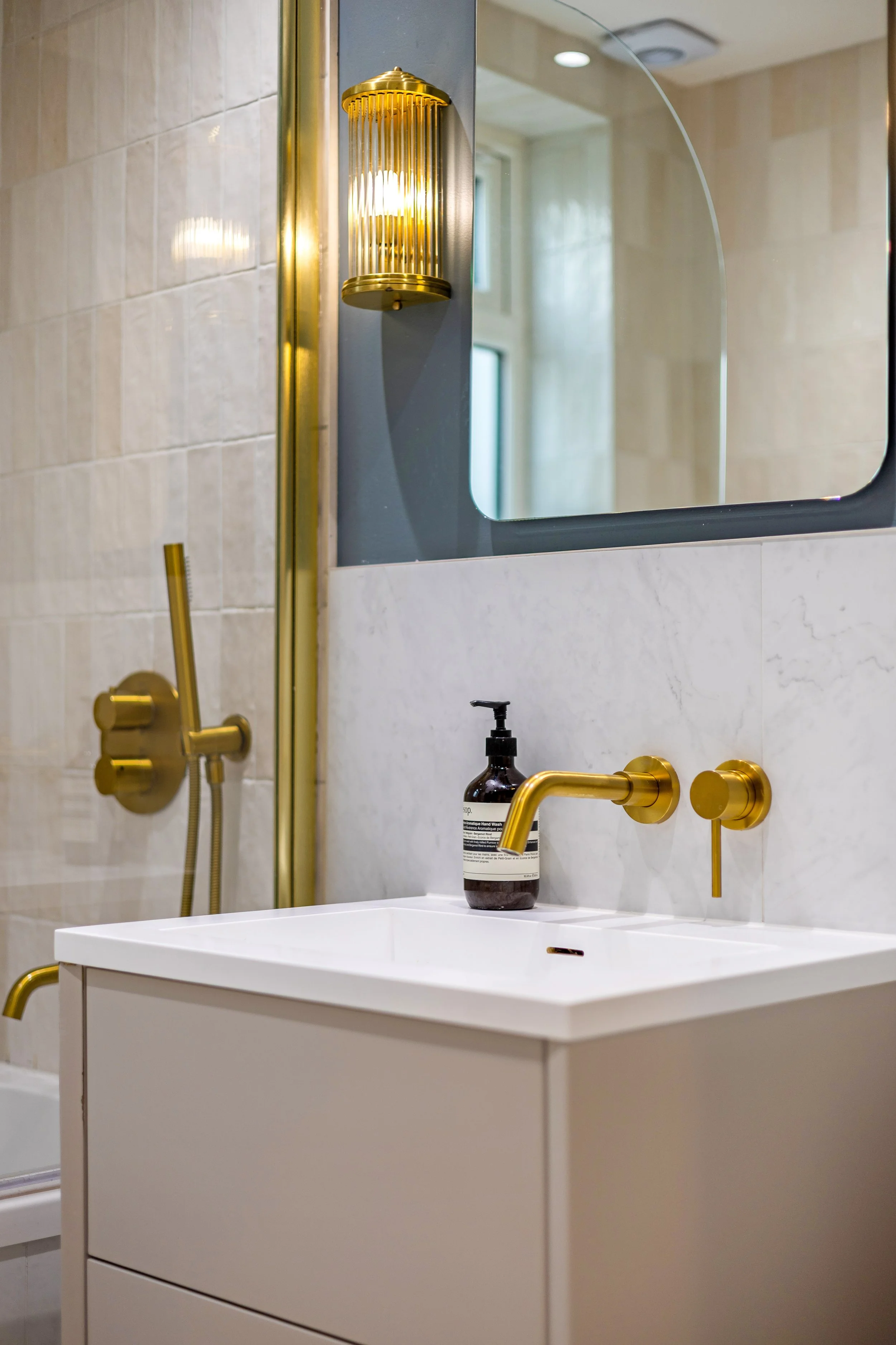 A modern bathroom mirror with gold fixtures, a white sink, a black soap dispenser, and a gold wall-mounted sconce with a frosted glass cover. The background shows a tiled wall, a shower with a gold showerhead, and a window reflected in the mirror.