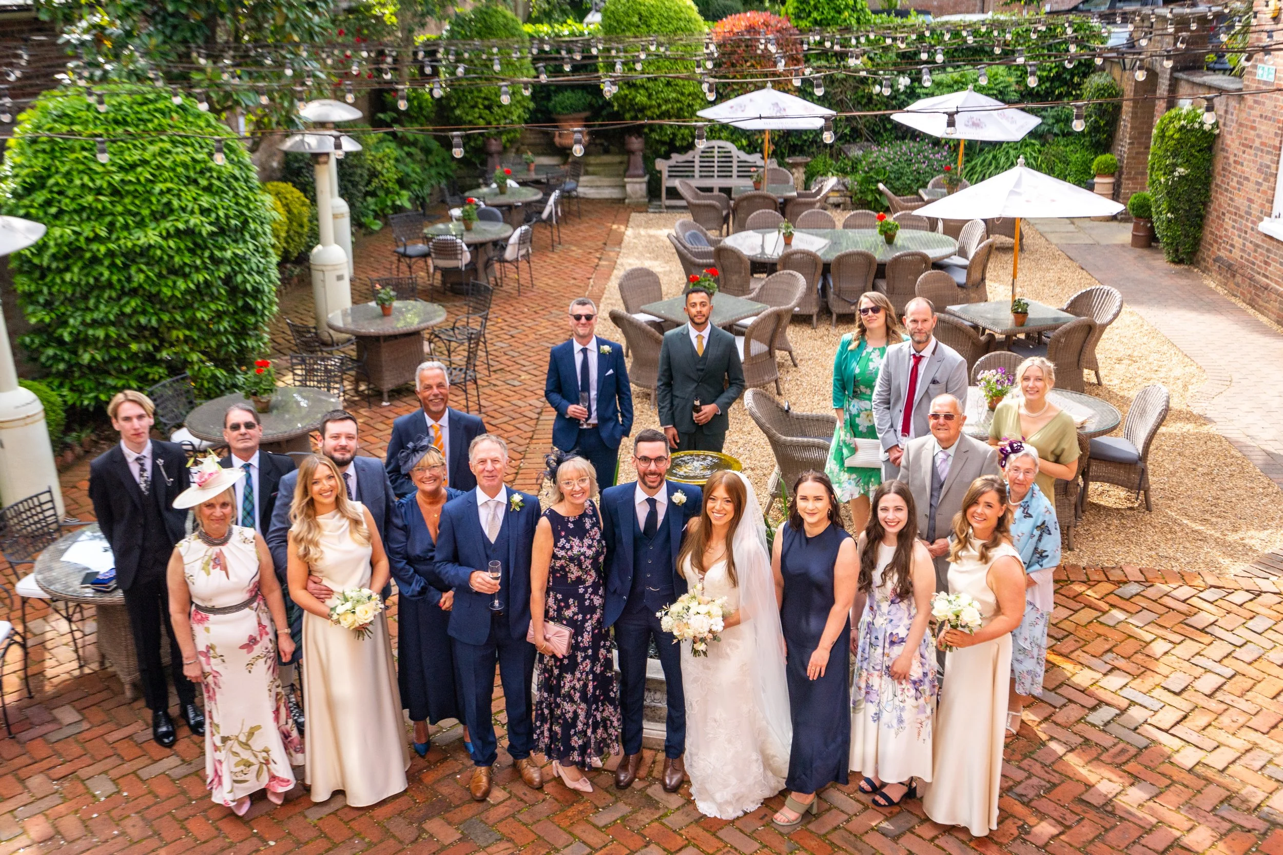 A group photo of wedding guests and the bride and groom standing on a brick patio at an outdoor wedding reception. There are tables with umbrellas and potted flowers surrounding the area, with greenery and brick walls in the background.