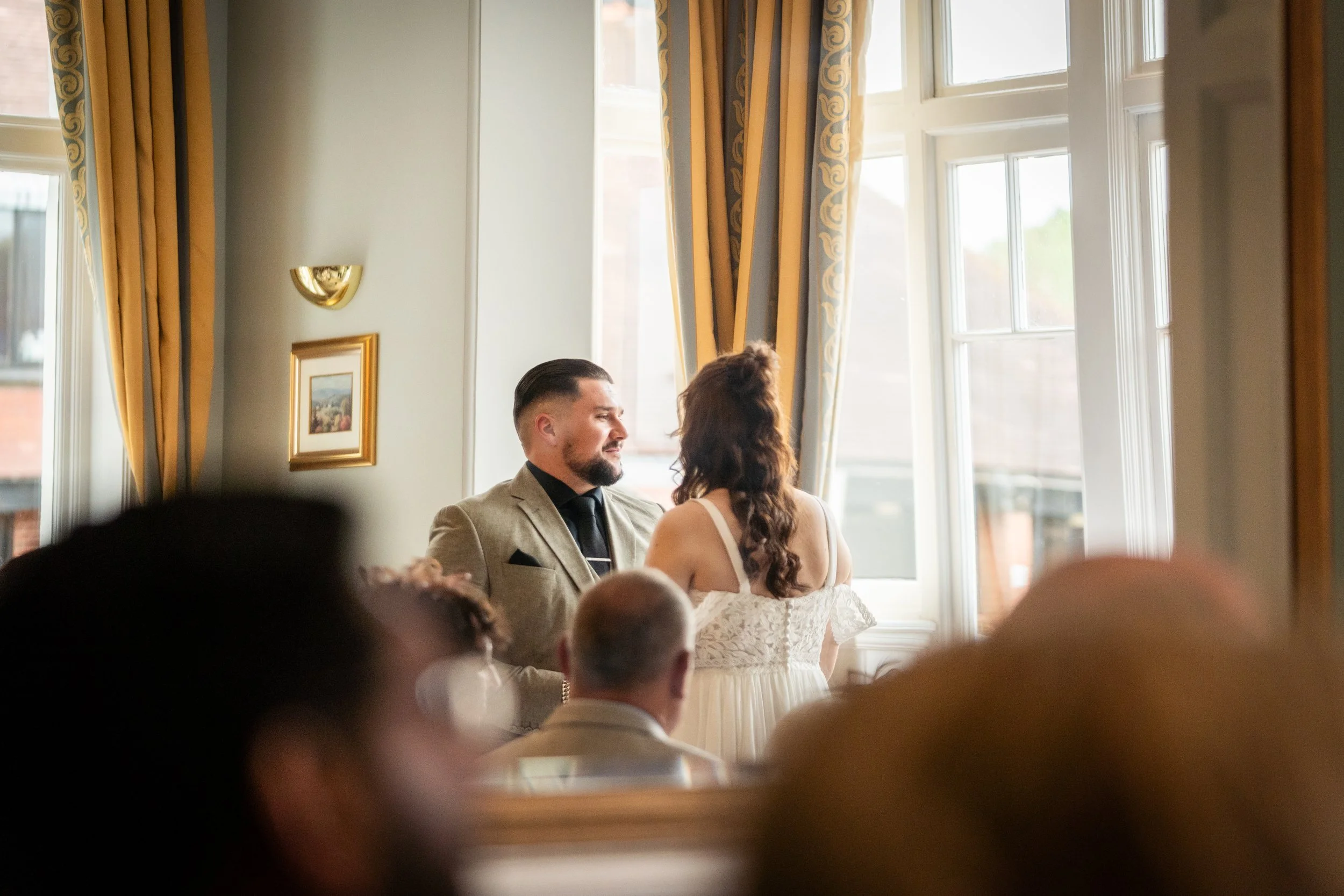 A couple getting married, facing each other in front of a gathered audience in a room with large windows and golden curtains.