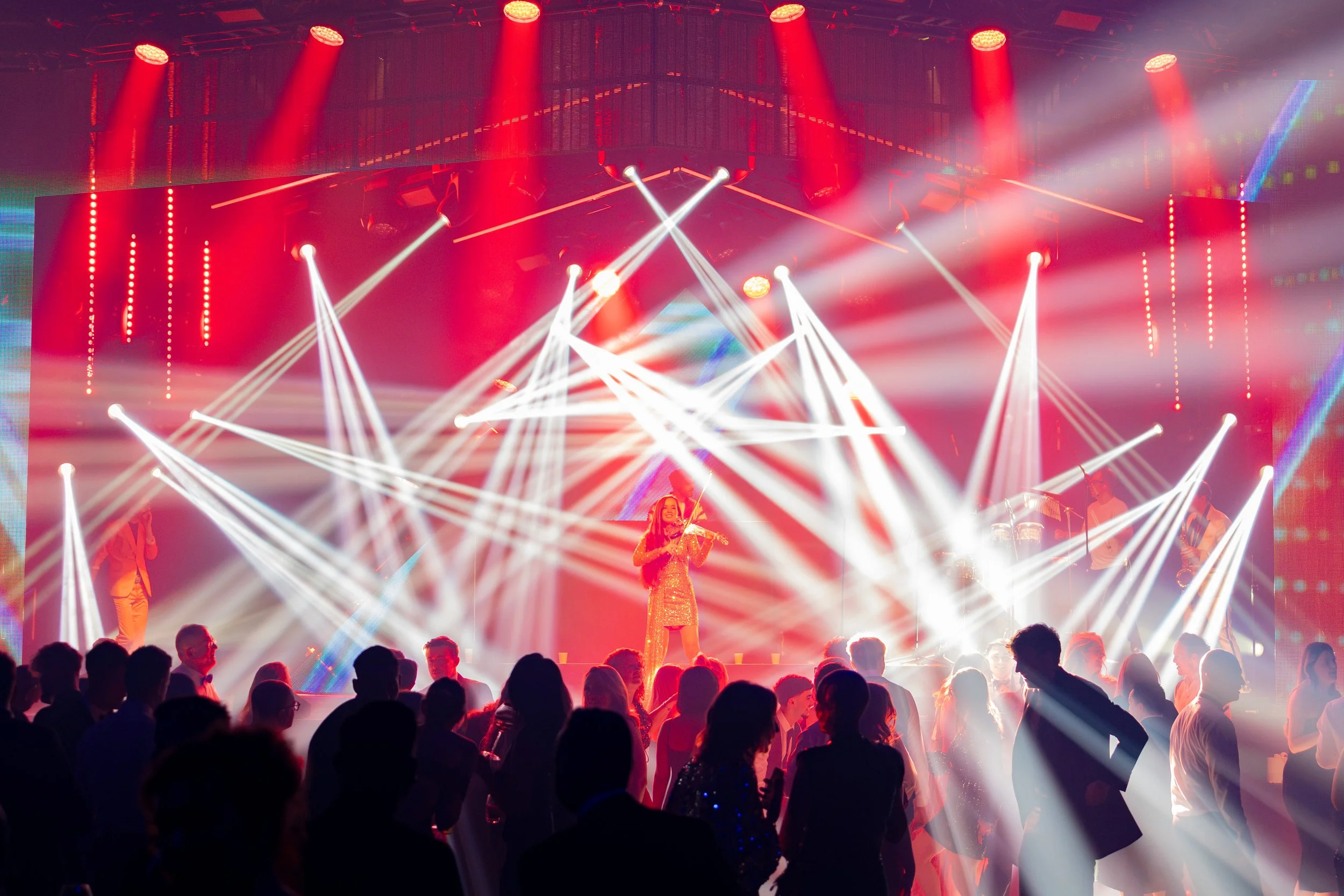 A lively concert scene with colorful lights, including red and white beams, illuminating a stage. A singer in a shiny gold dress performs with a band, while the audience watches and enjoys the show.
