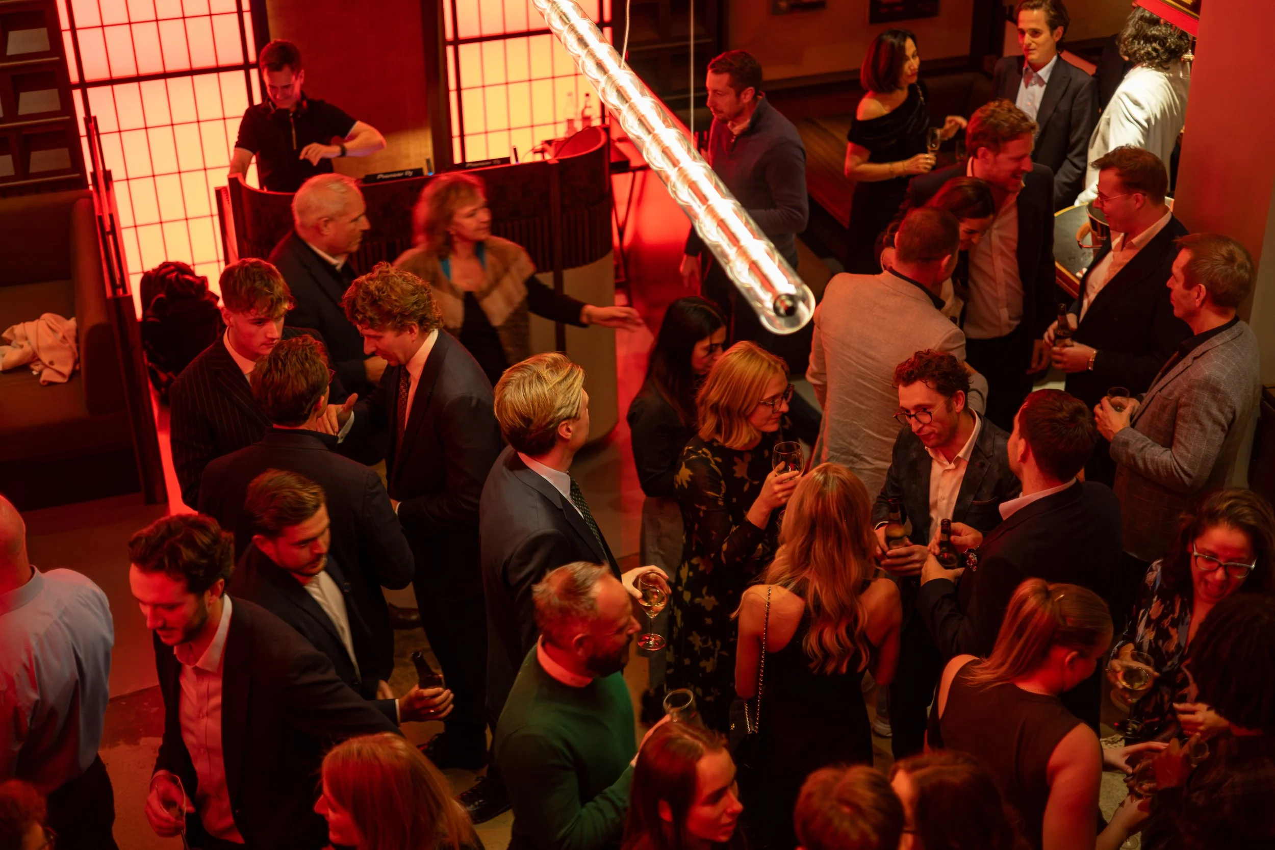 A crowded social gathering with people engaged in conversation and holding drinks, illuminated by red lighting in an indoor venue.