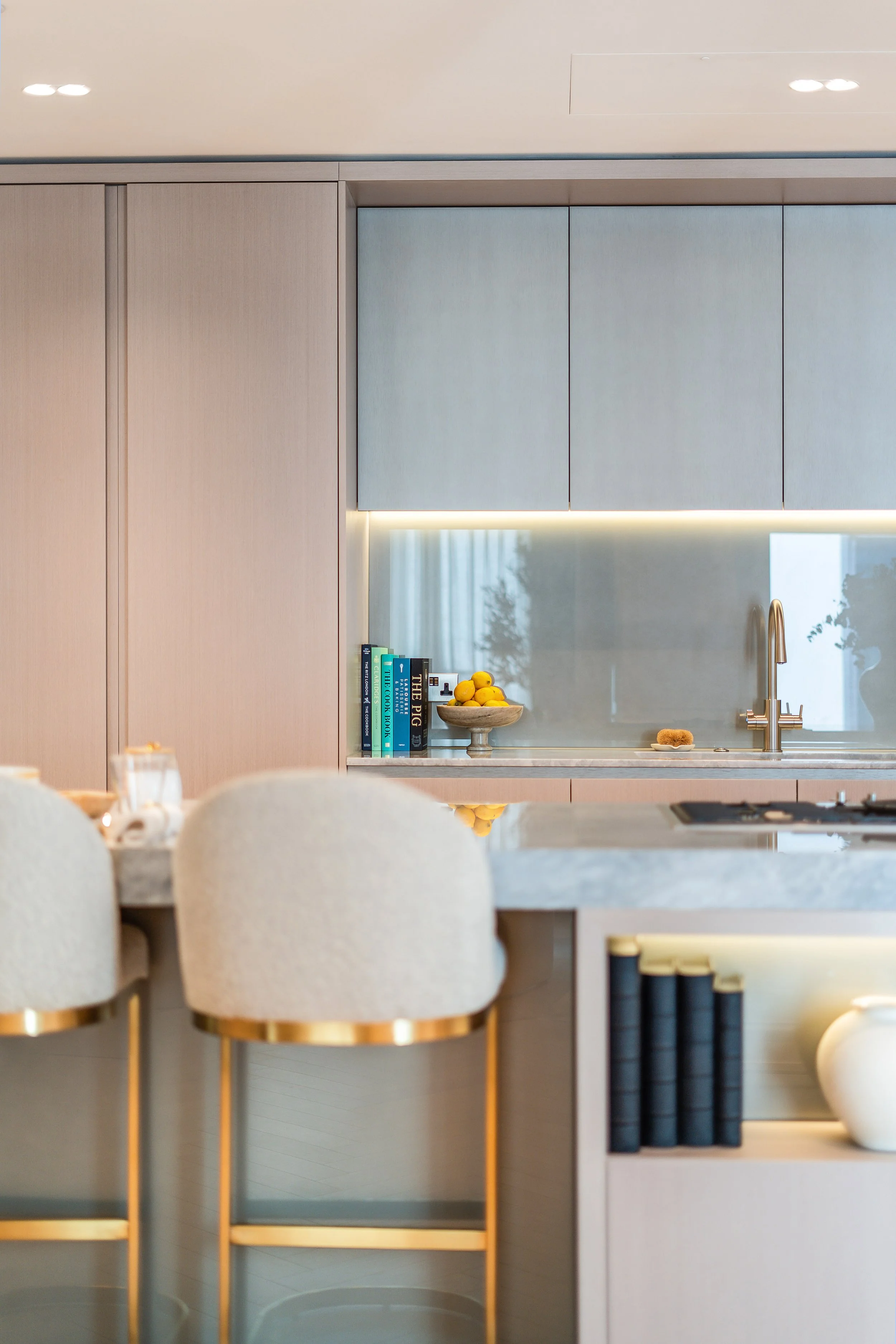 A modern kitchen with beige cabinetry, a sleek white countertop, and a backsplash with under-cabinet lighting. A bowl of lemons and a few books are on the counter near a stainless steel faucet and sink. The reflection of windows and trees can be seen in the backsplash.