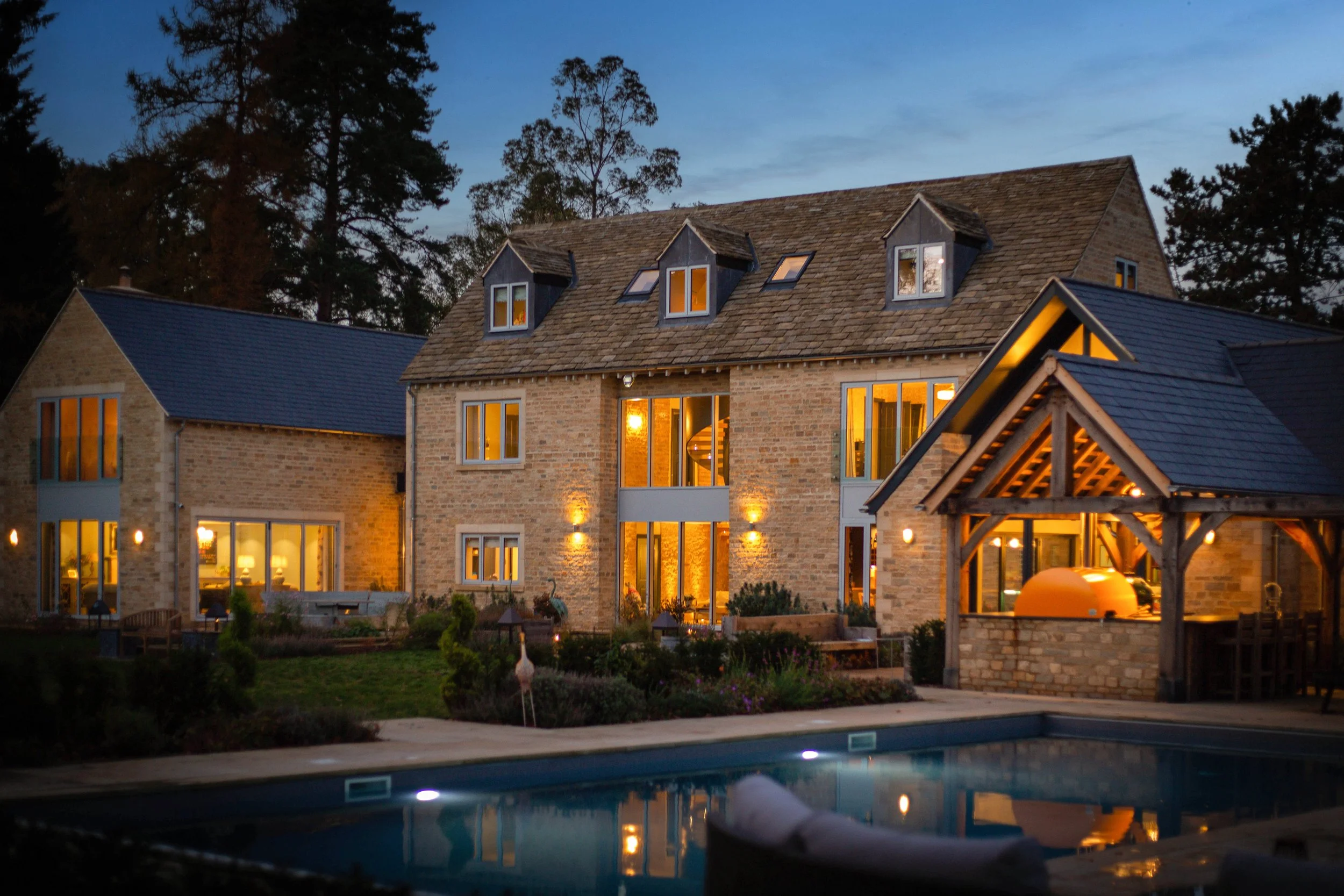 A large brick house with a steep shingled roof and multiple dormer windows illuminated from within, surrounded by tall trees at dusk, with a swimming pool in the foreground.