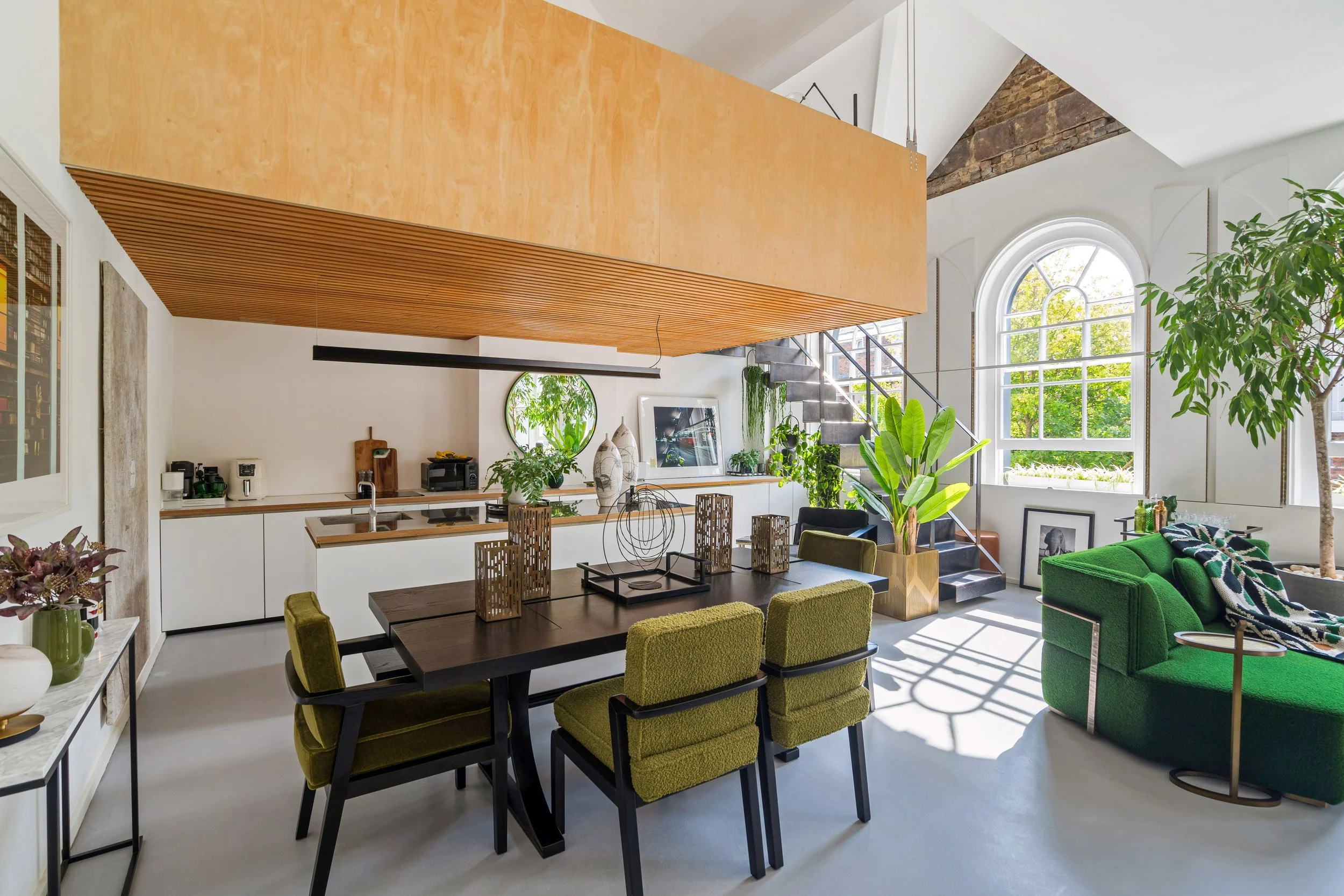 Bright and spacious living room with large arched window, green plants, green sofa, black staircase, wooden dining table with green chairs, and modern decor elements.