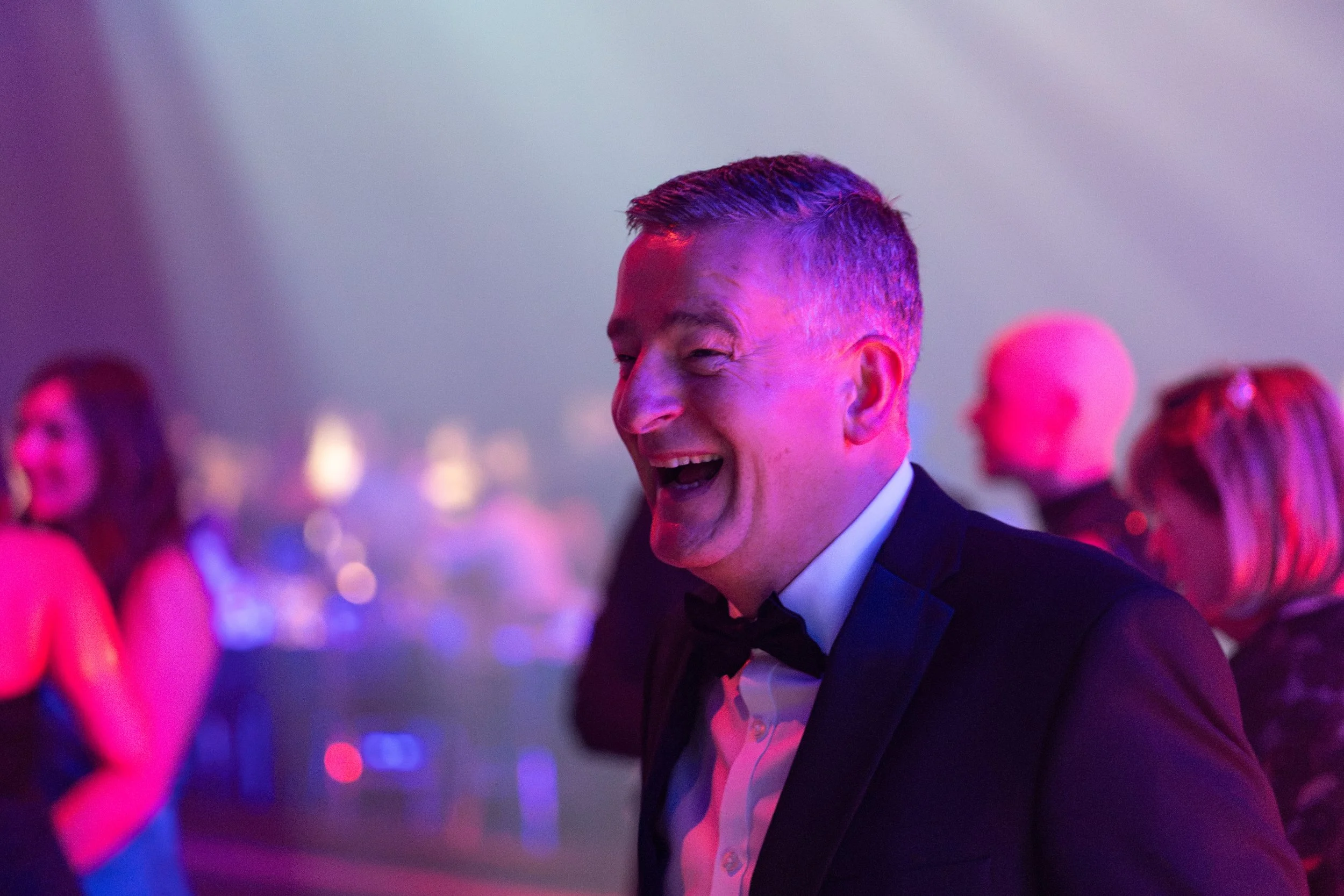 A man in a tuxedo laughing and dancing at a party or celebration, with pink and purple lighting, surrounded by other women in fancy dresses.