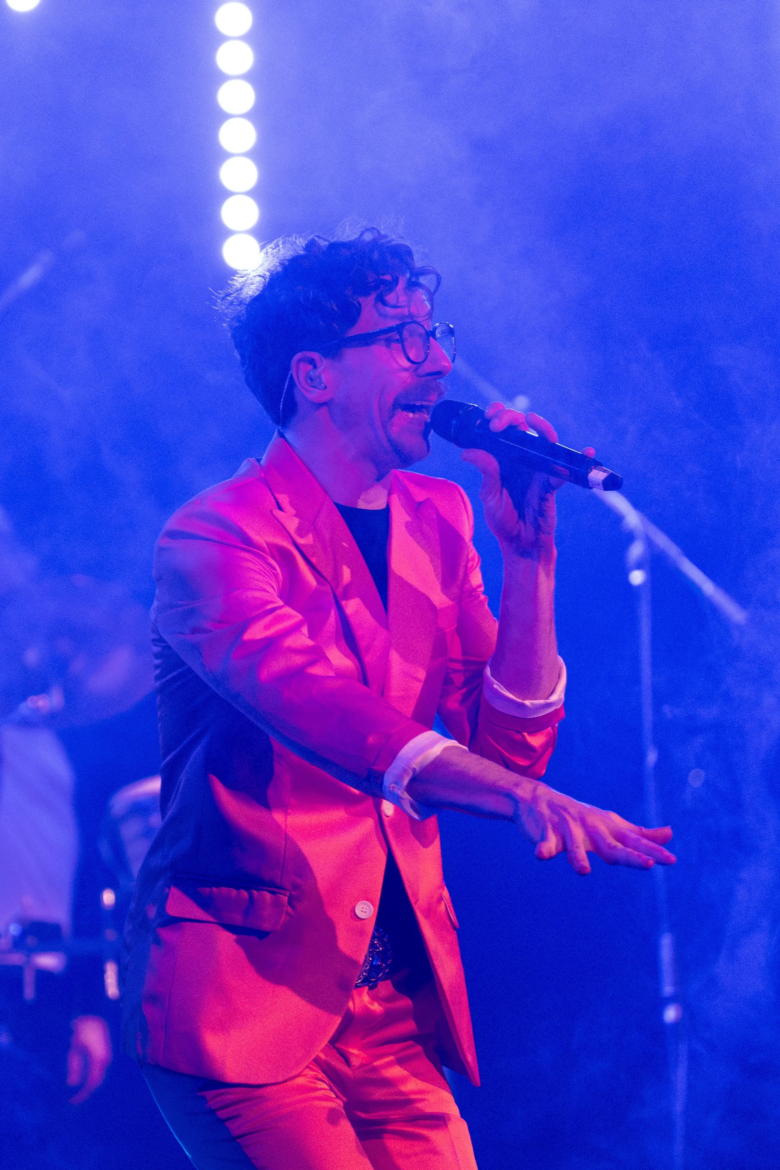 A man with curly hair and glasses, dressed in a pink suit, singing into a microphone on stage with blue and purple lighting.