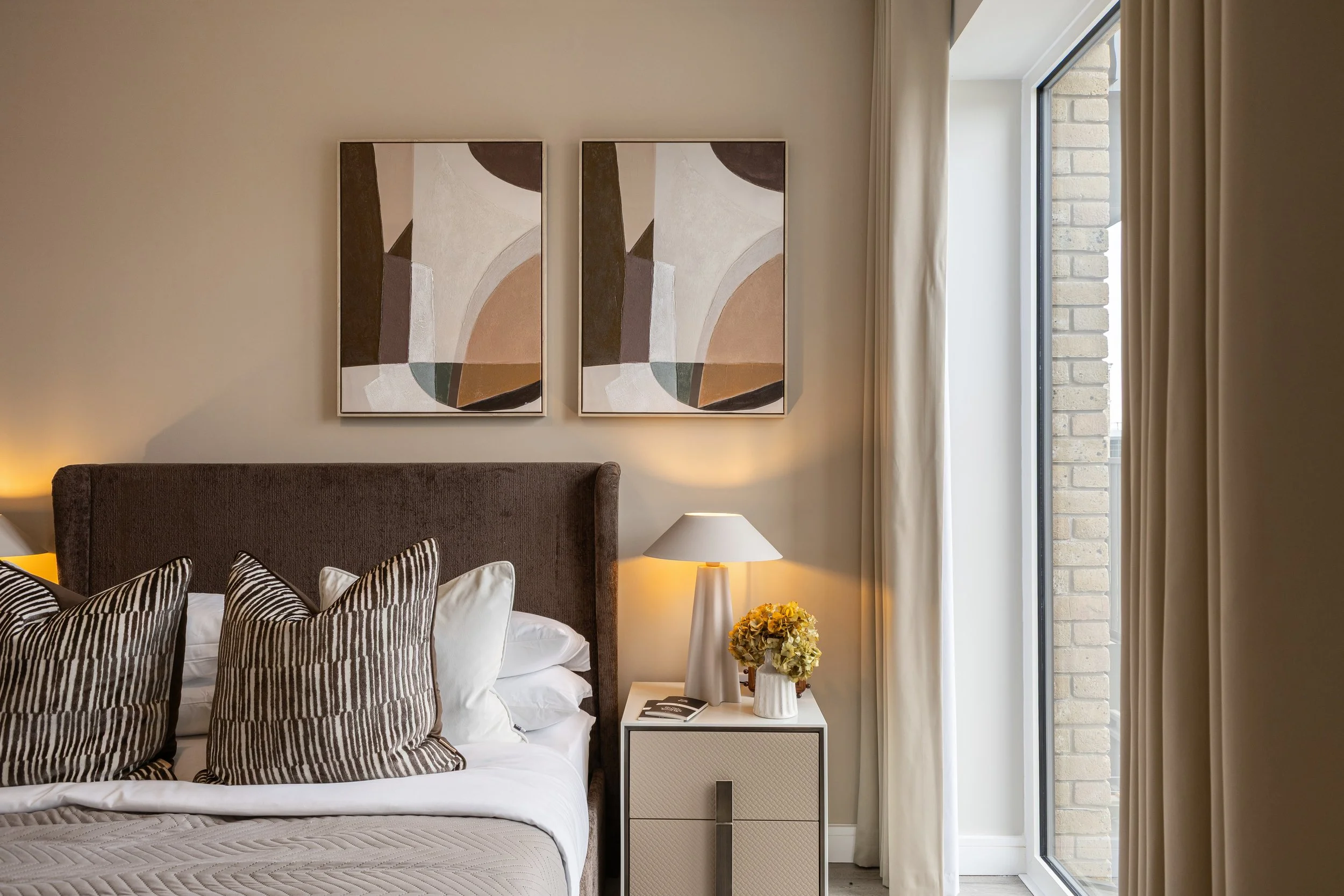 A bedroom with a bed, multiple zebra-patterned pillows, a nightstand with a lamp, and abstract artwork on the wall. There is a window with curtains and a brick exterior wall visible outside.