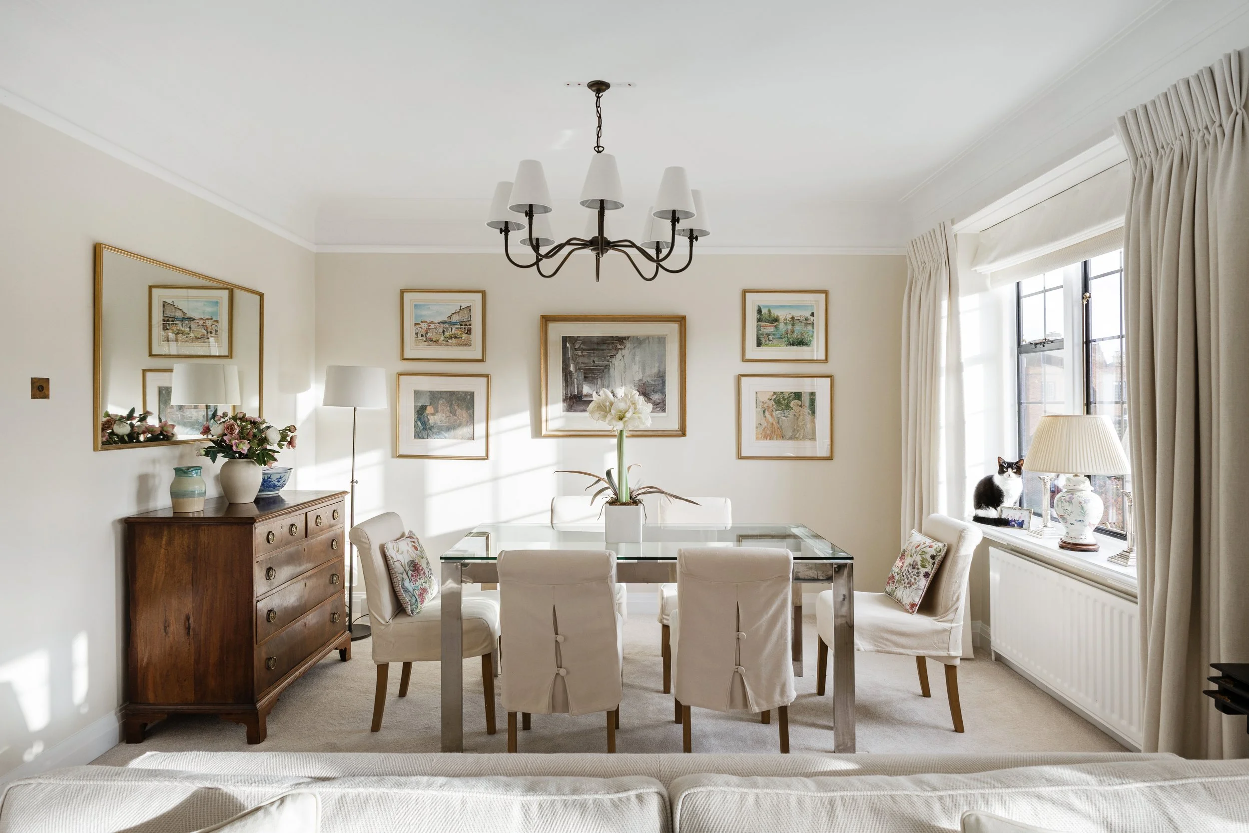 A living room with beige walls, decorated with framed artwork, a wooden sideboard with vases and flowers, a glass dining table with white chairs, a window with curtains, a chandelier, and a black-and-white cat sitting by a lamp on a side table.