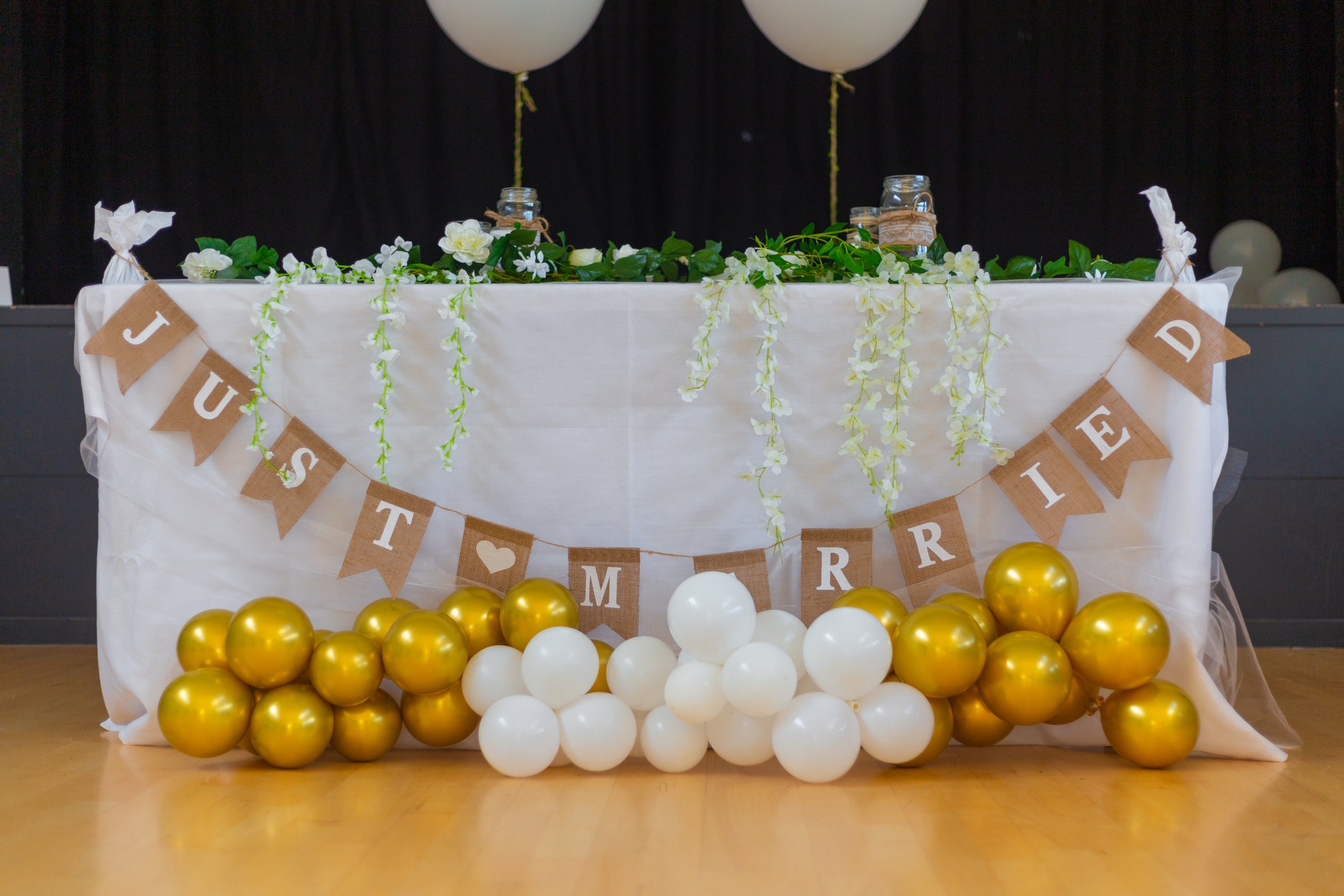A decorated table with a white tablecloth, green foliage, white flowers, and jars on top. A banner with burlap flags spells 'JUST MARRIED' with a heart in the middle, hanging in front. Gold and white balloons surround the table at the base. Two large white balloons are attached behind the table, with a black backdrop.