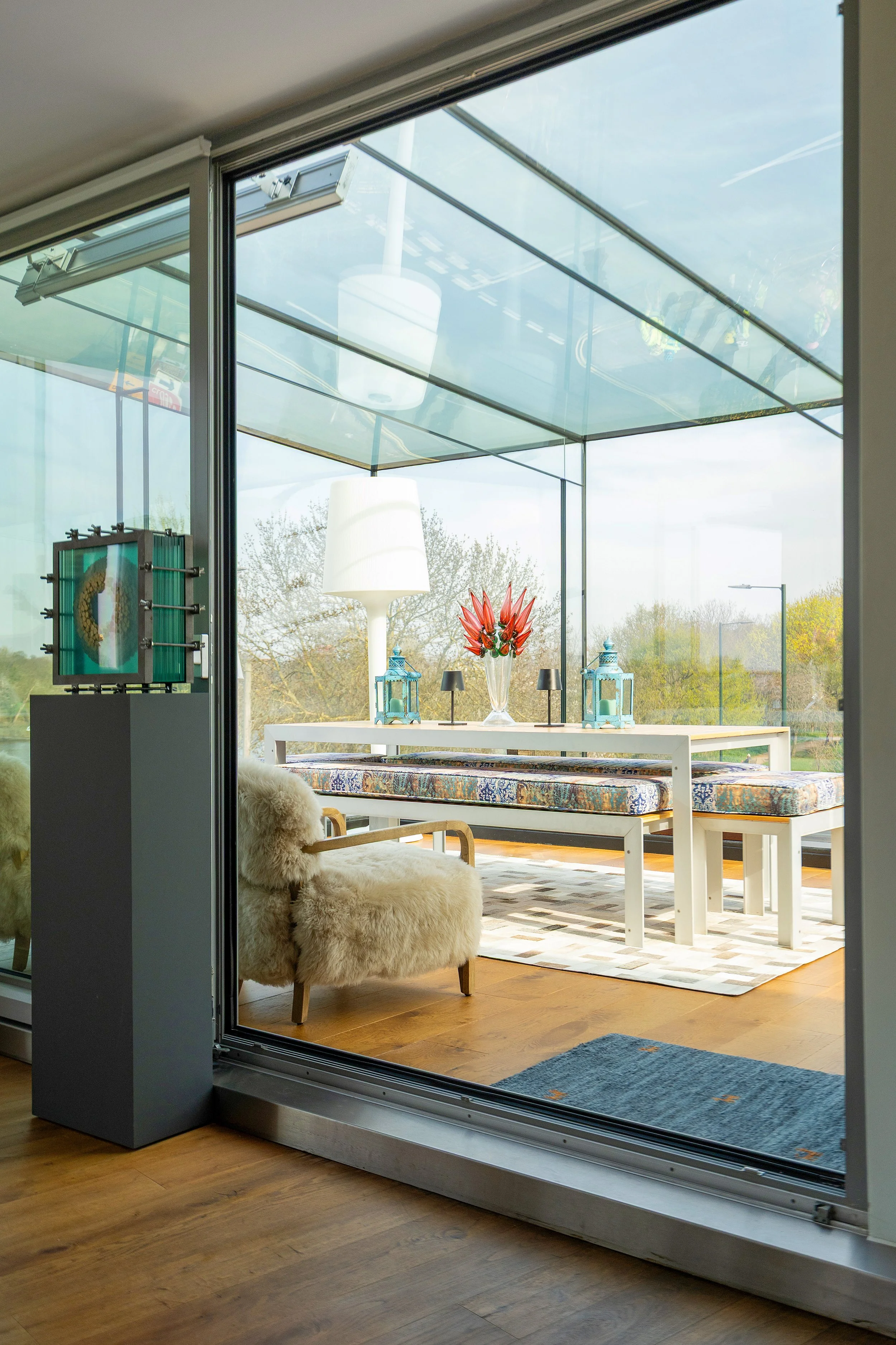 Interior view through glass sliding door showing a living space with a cozy armchair covered in sheepskin, a white table with red flowers, and a view of outdoor trees and sky.