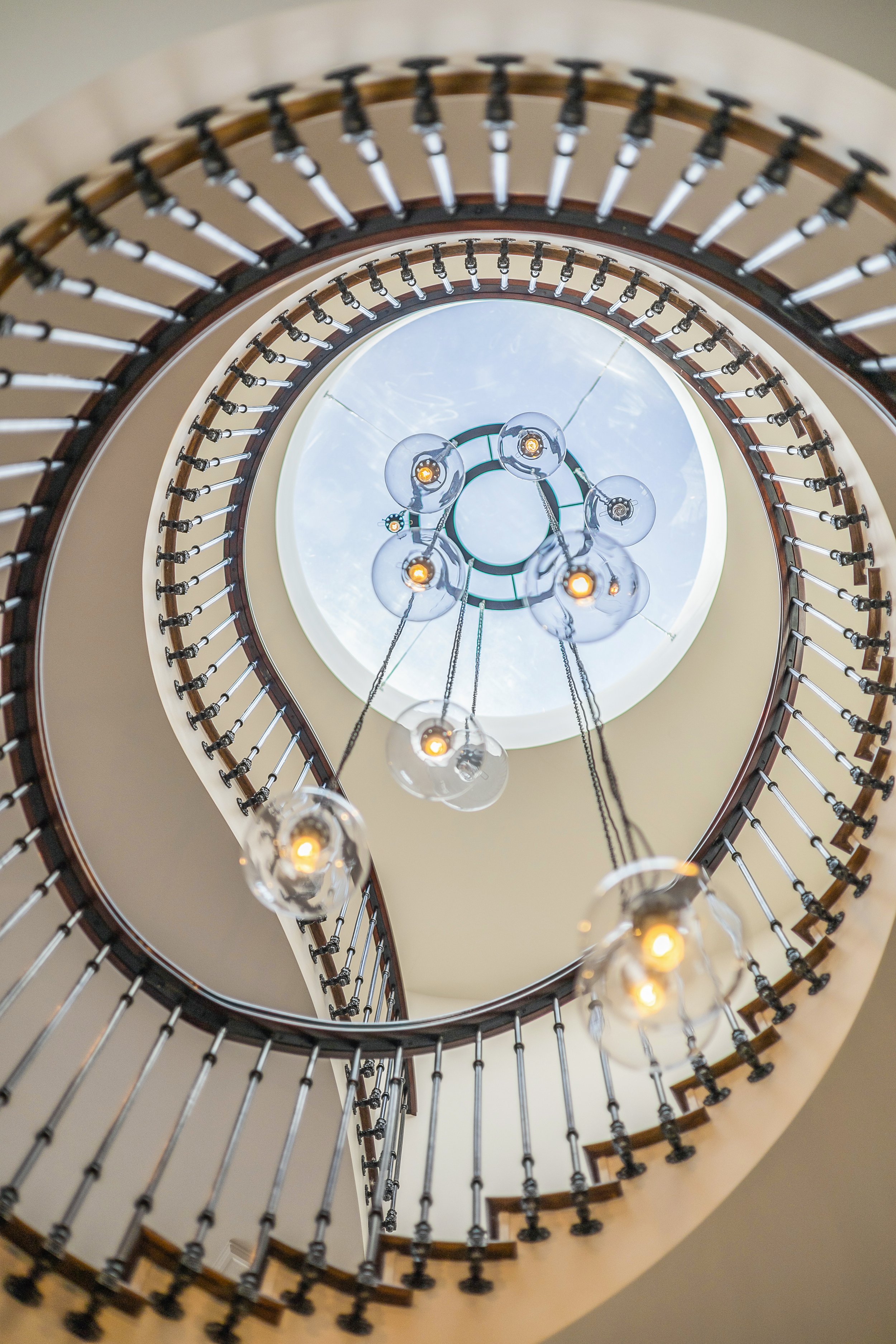 A spiral staircase viewed from the bottom, with hanging pendant lights and a circular skylight at the top.