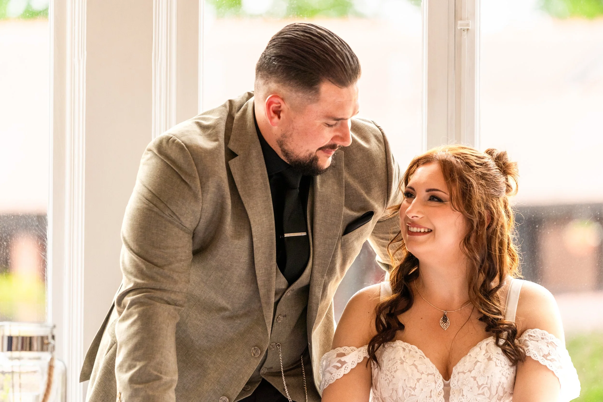 A man in a beige suit leaning towards a woman in a white lace dress, both smiling, indoor setting with large windows.